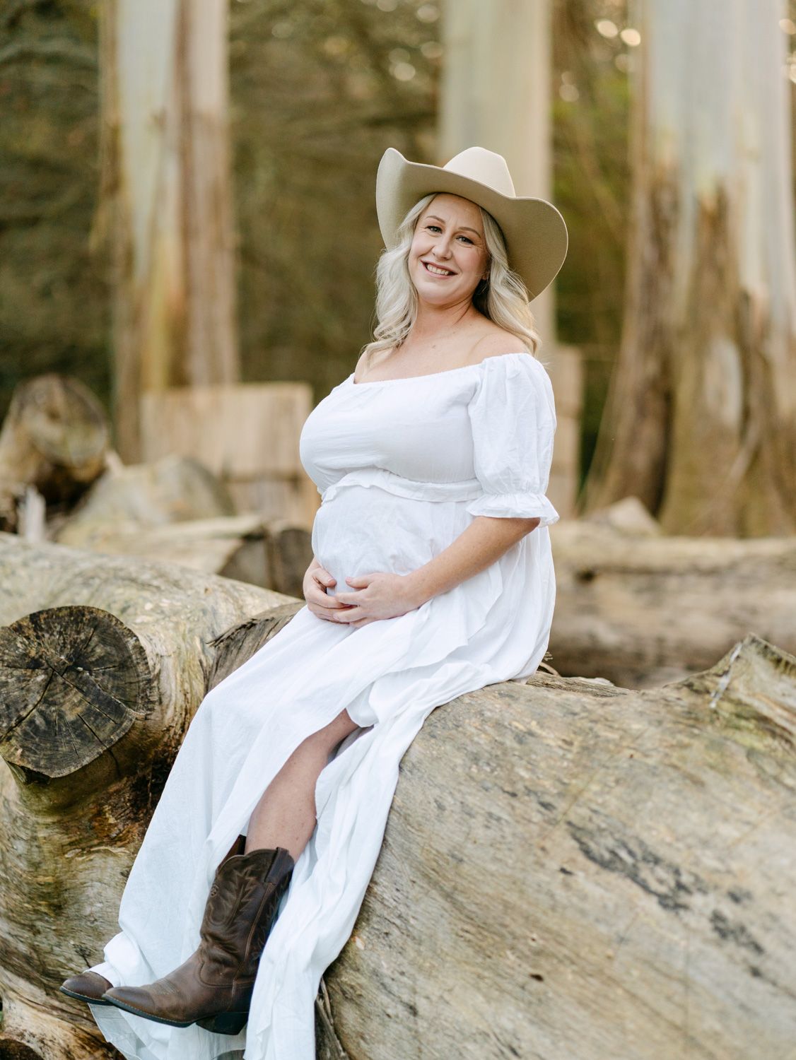 Pregnant woman in white off-shoulder dress, cowboy hat, and brown boots sitting on logs outdoors.