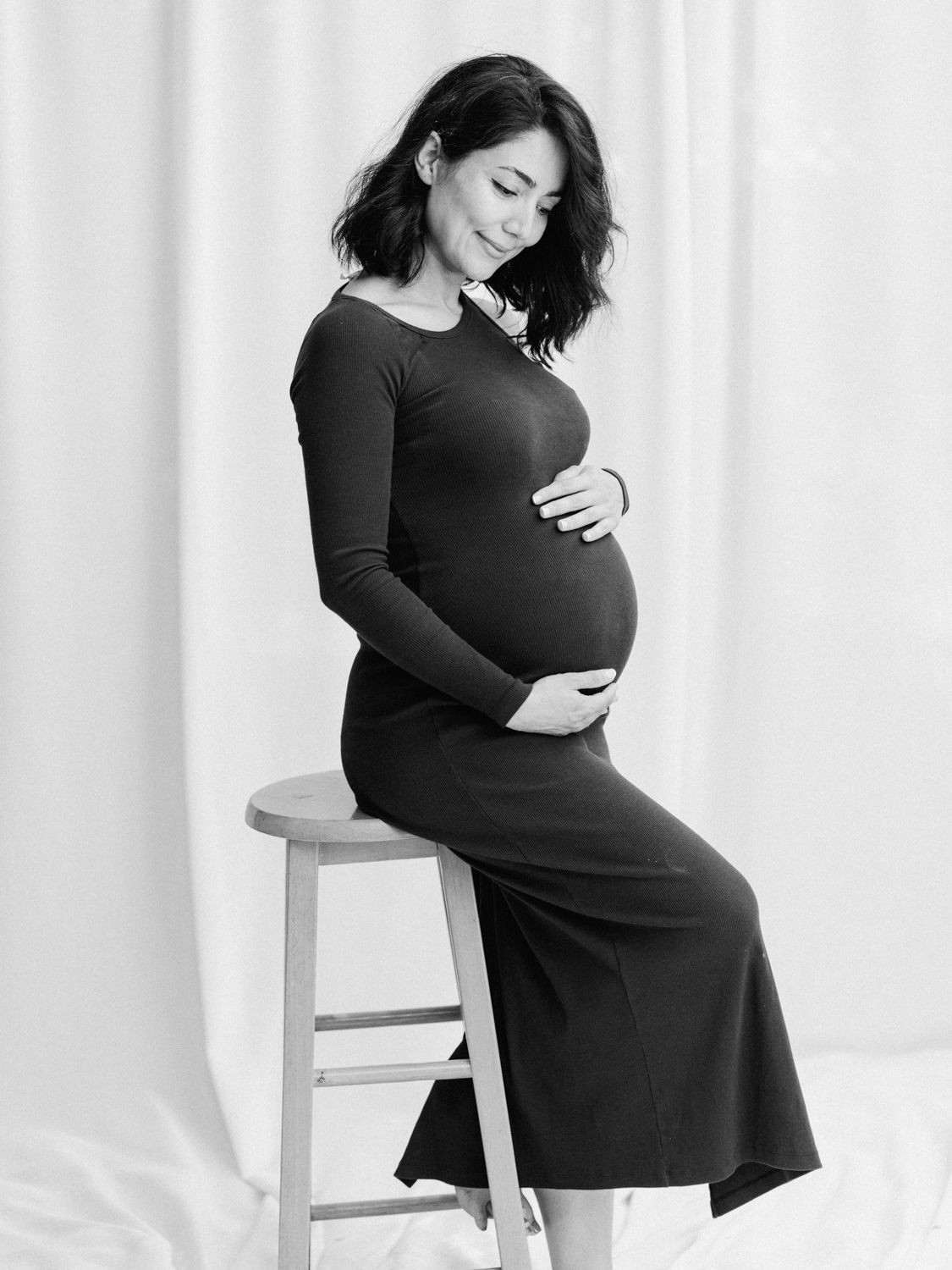 Black and white maternity photo of a pregnant woman in a dark dress sitting on a stool, cradling her baby bump.
