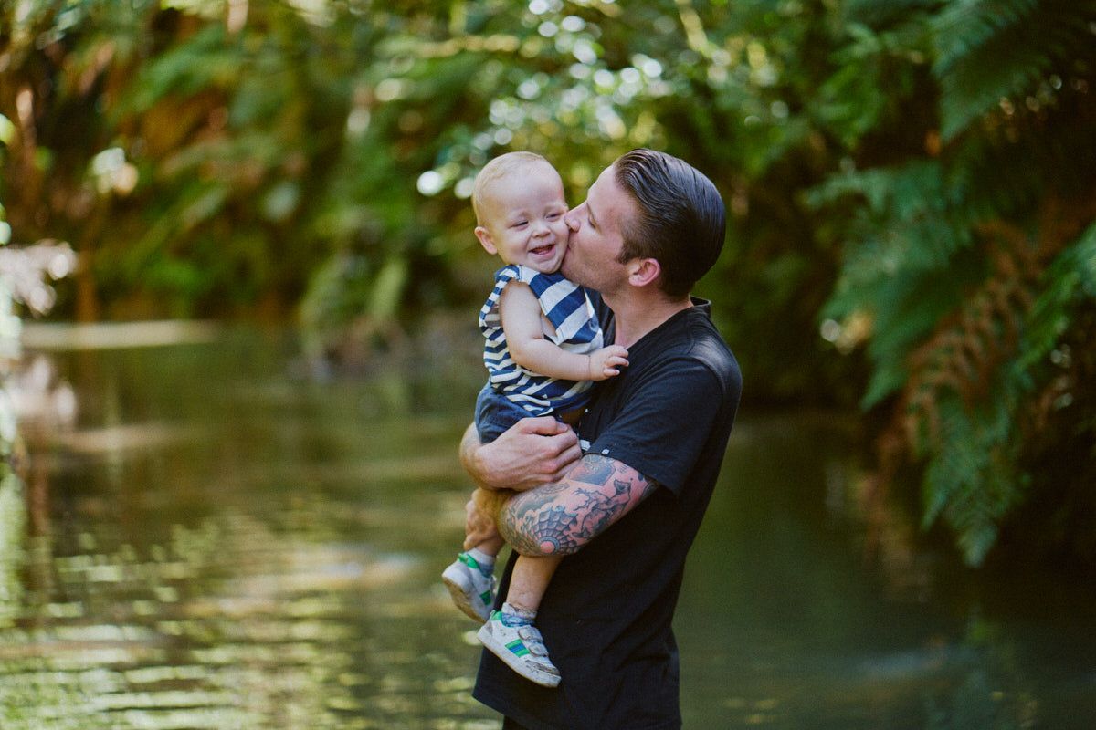 A tattooed man lovingly holds a laughing baby outdoors near a serene green pond surrounded by lush ferns.
