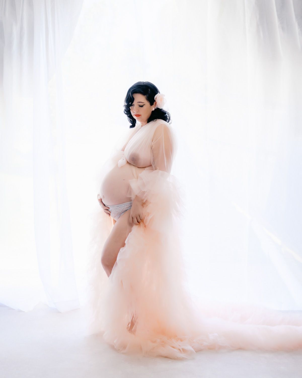 Pregnant woman in sheer blush pink tulle robe cradling baby bump, posed against soft white curtains.