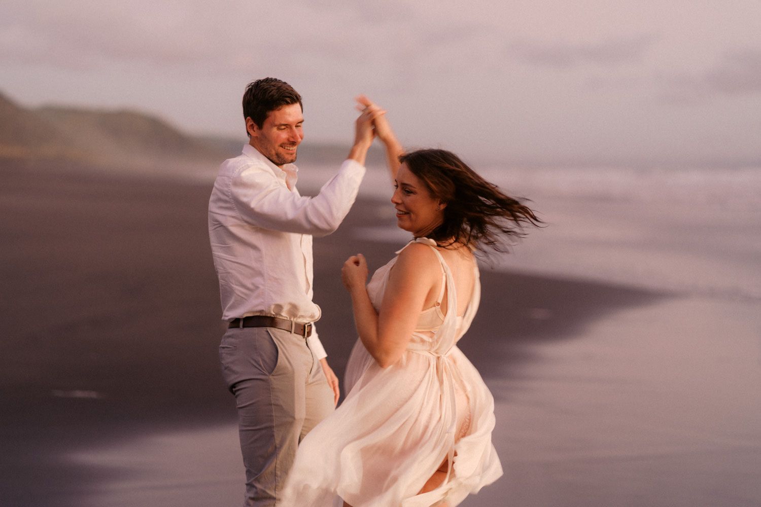Couple dancing romantically on a misty beach at sunset, woman in flowing pink dress, man in white shirt.