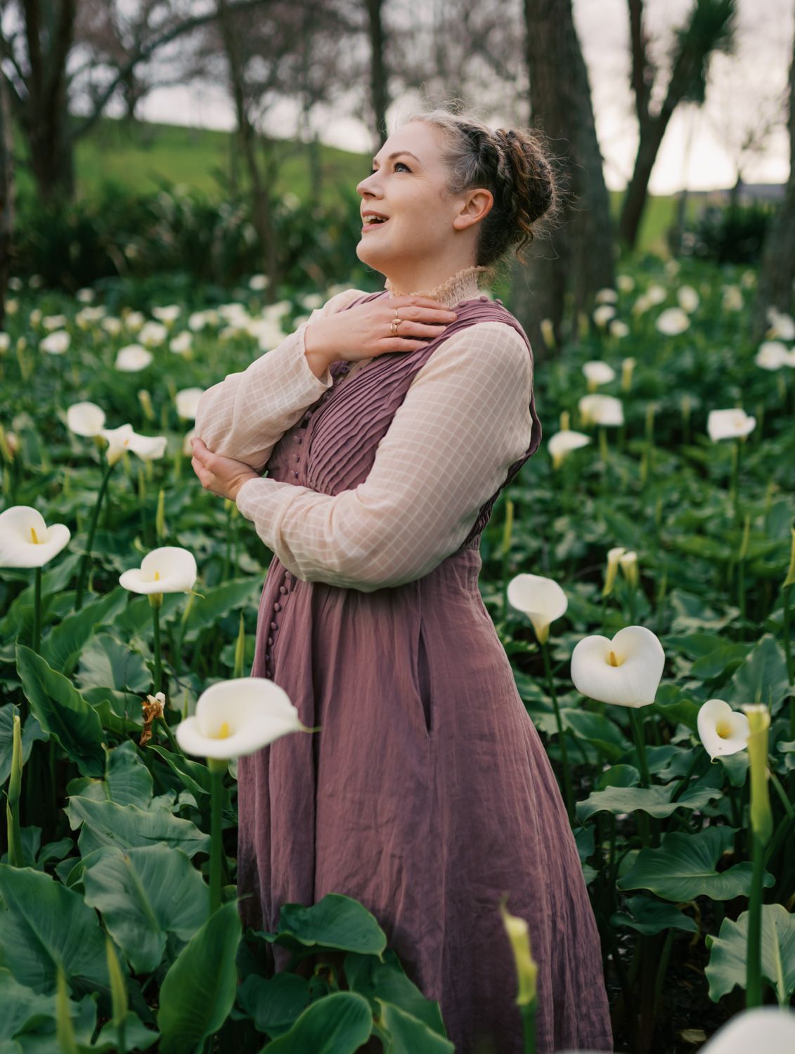 Woman in mauve dress standing among white calla lilies in a lush garden, looking upward peacefully.