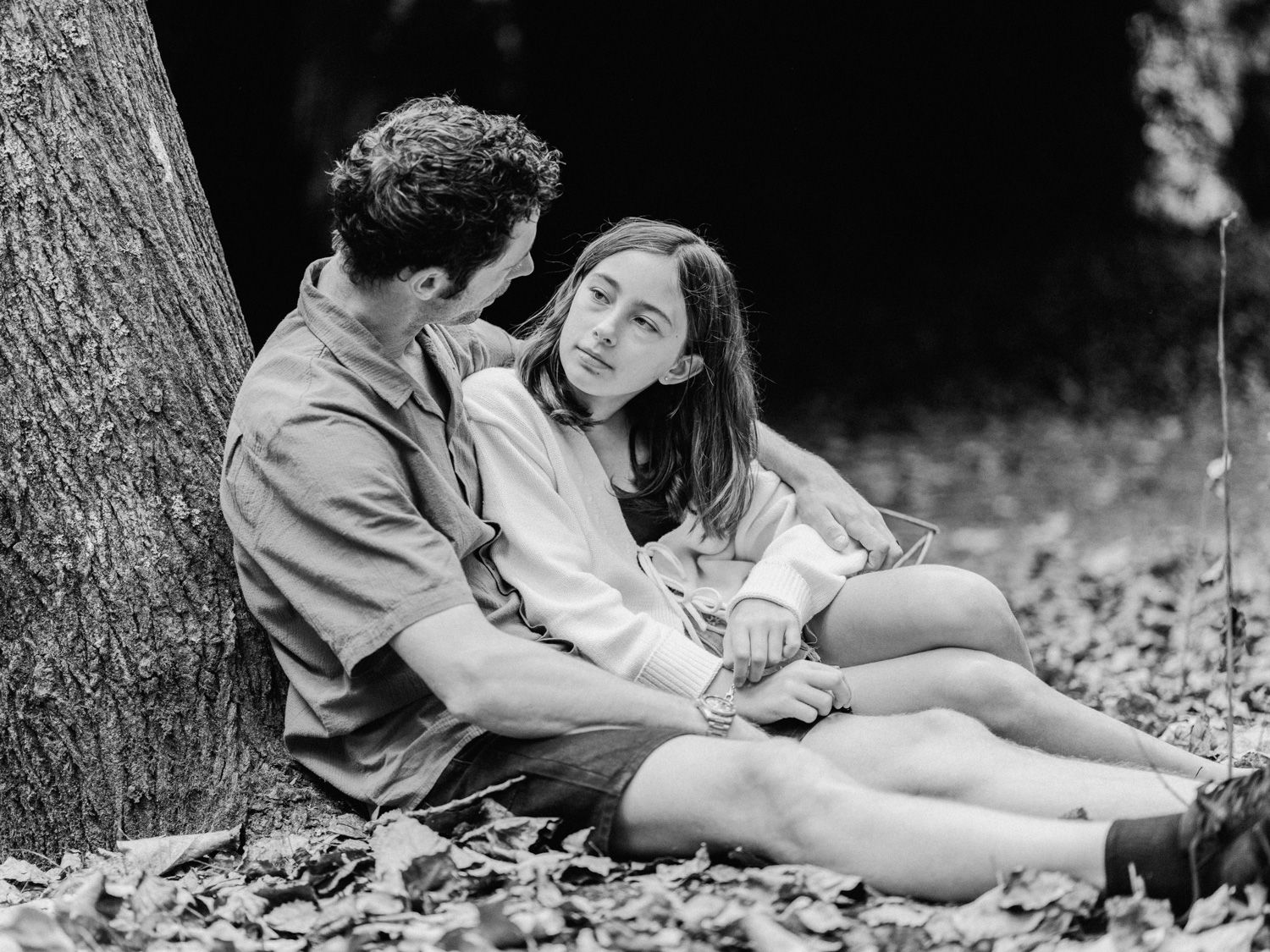 Black and white photo of a couple sitting close together against a tree, sharing an intimate moment outdoors.