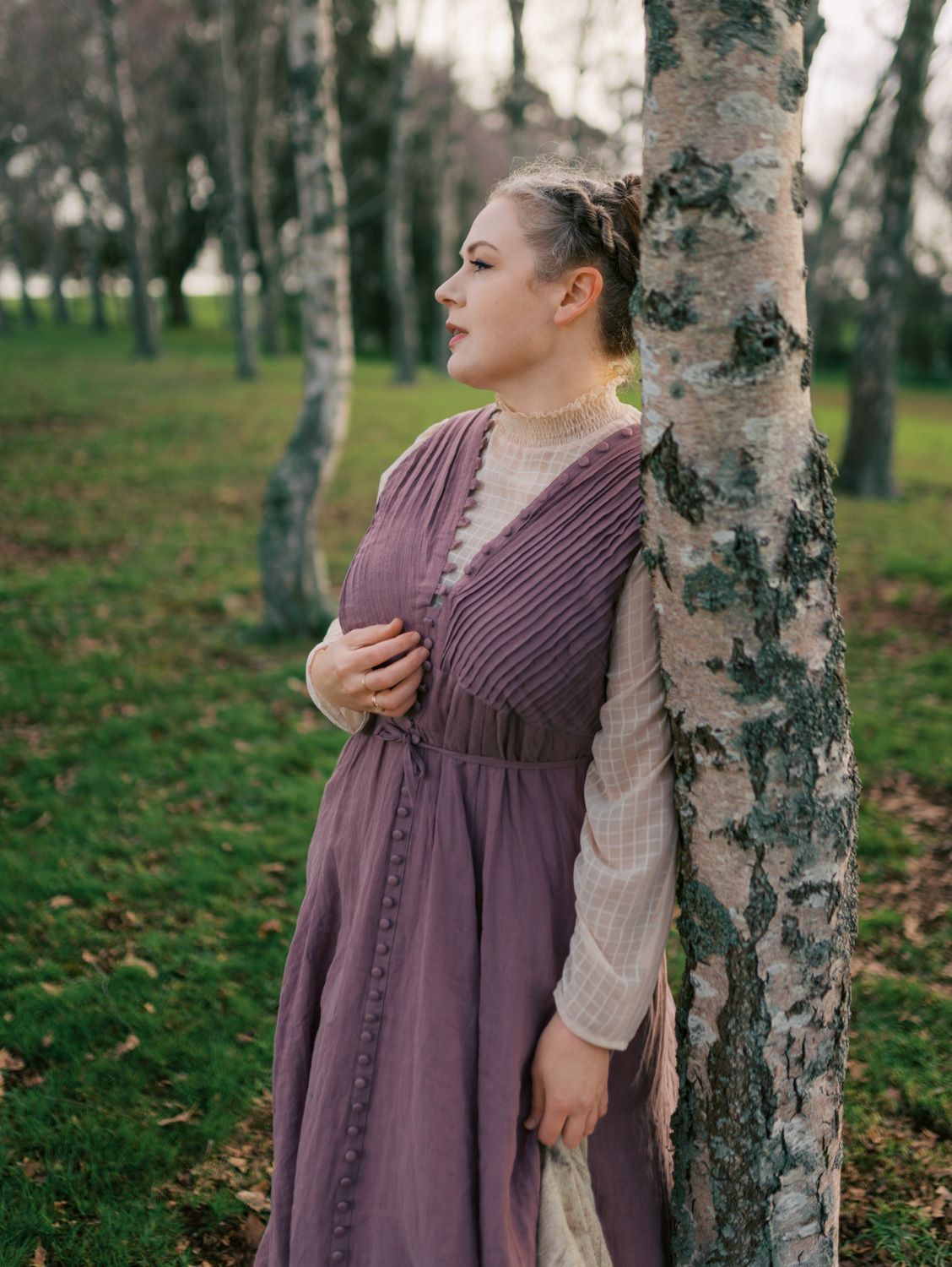 Woman in purple medieval dress leaning against birch tree in autumn park setting.