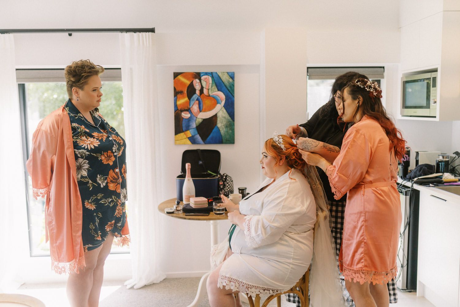 Bride getting hair styled by hairdresser while bridesmaid watches in bright room with colorful artwork.