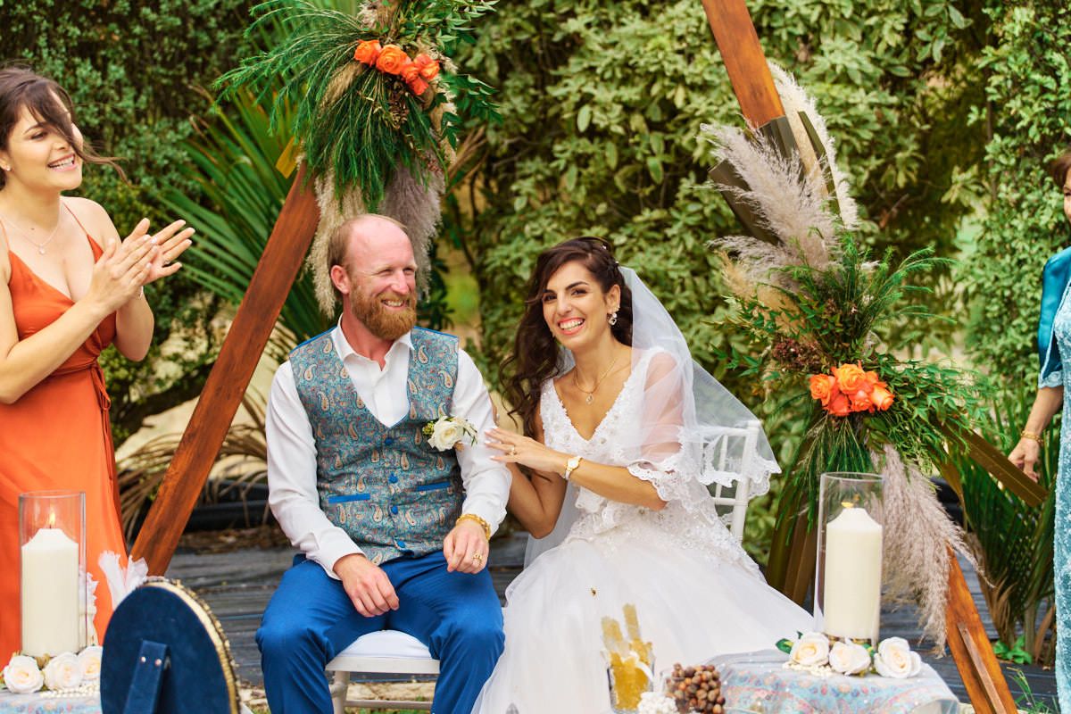 Happy couple laughing during outdoor boho wedding ceremony under triangular arch with tropical greenery and orange flowers.