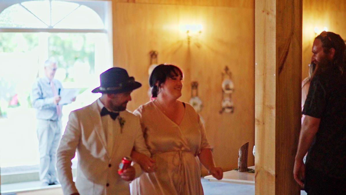 Couple in vintage attire dancing at a rustic indoor wedding reception with guests in background.