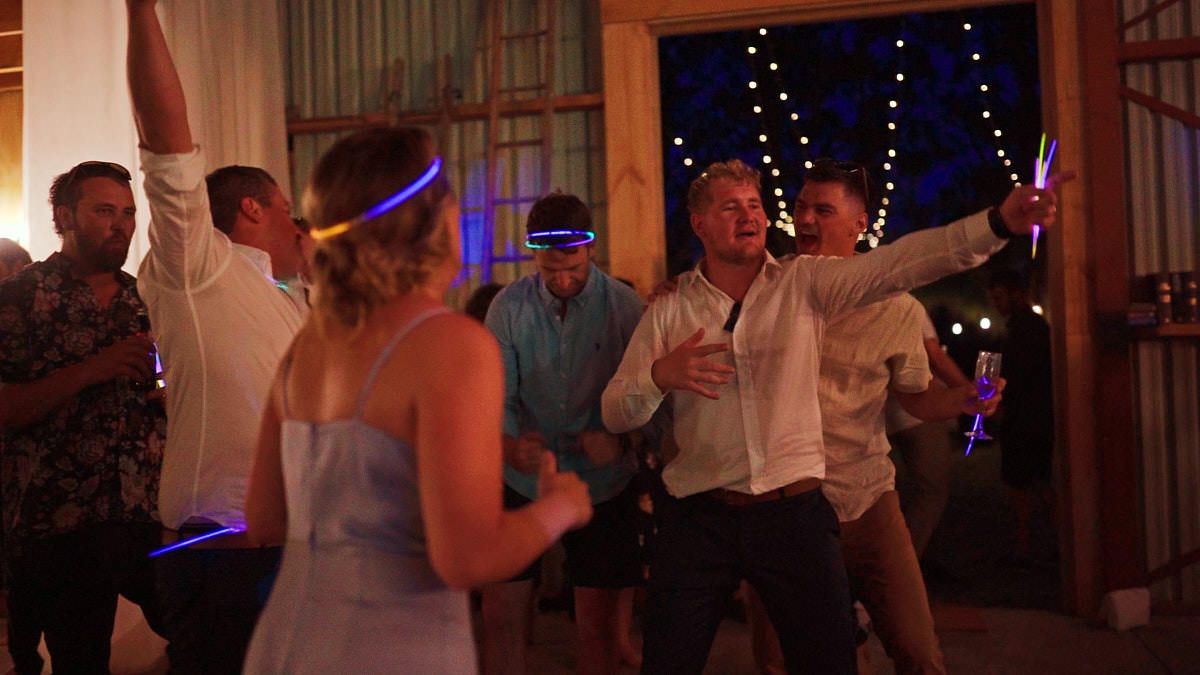 Wedding guests dancing with glow sticks at a lively rustic barn reception celebration at night.