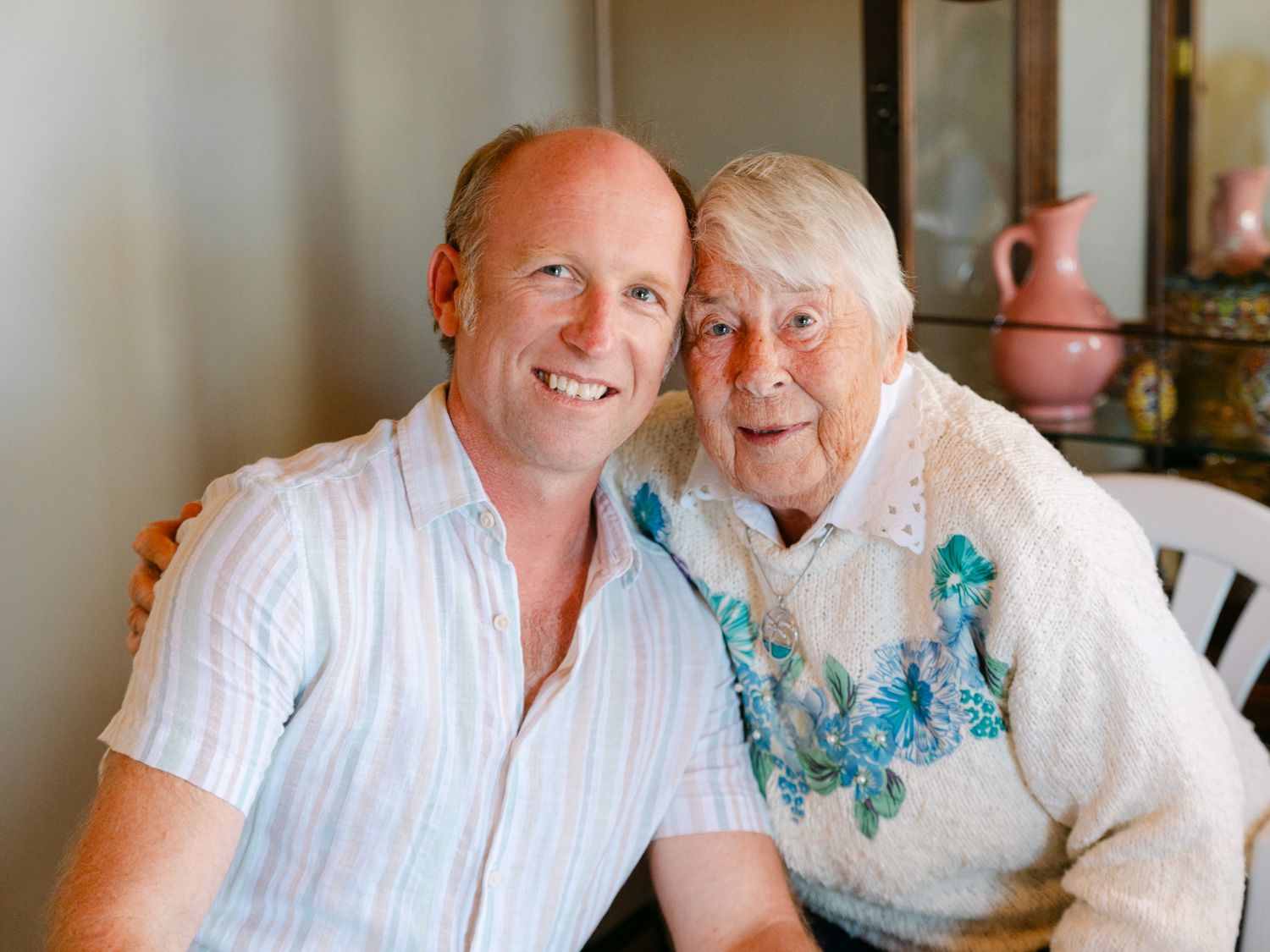 A smiling middle-aged man poses cheek-to-cheek with an elderly woman in a warm, cozy indoor setting.