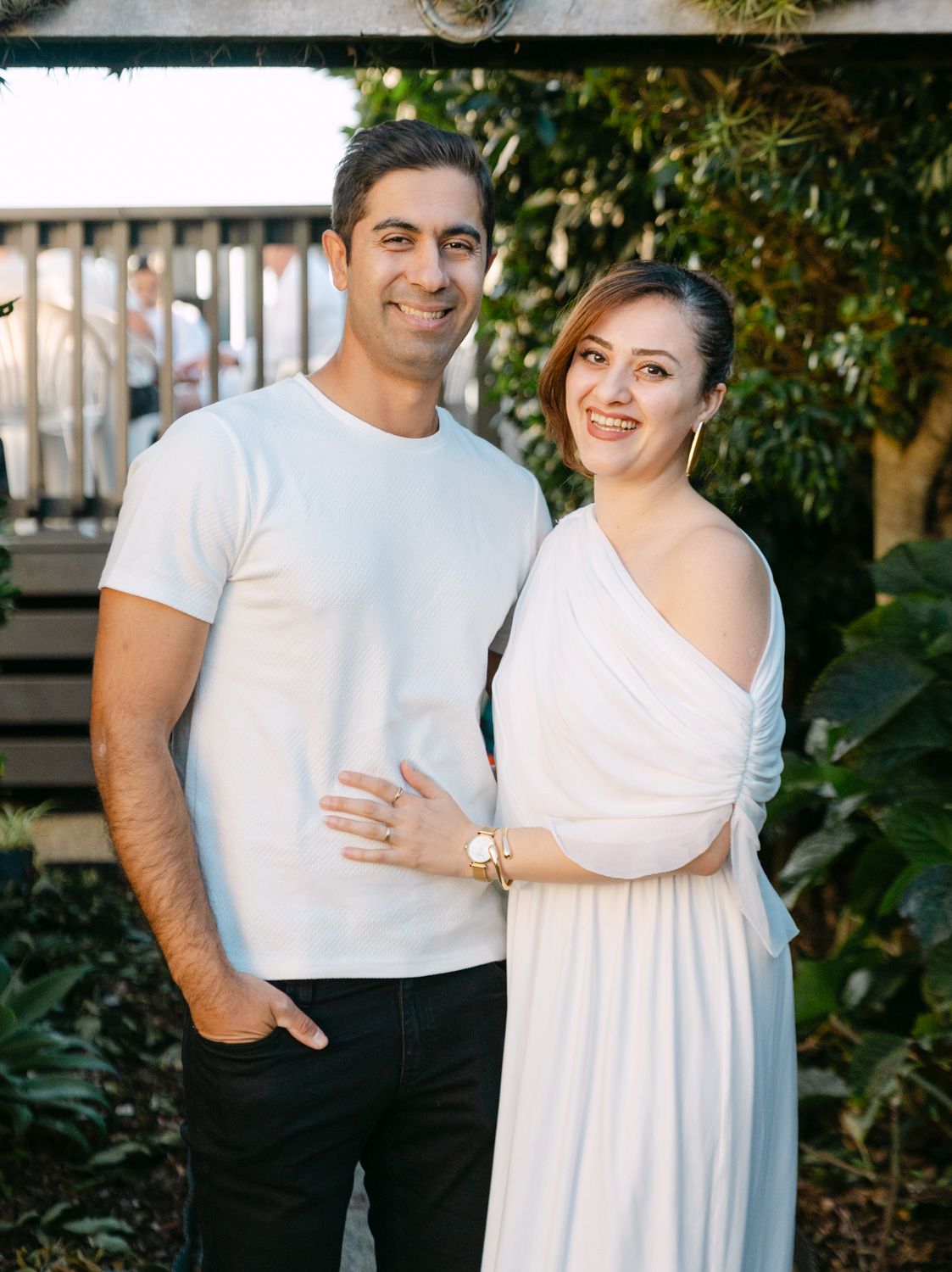 A smiling couple in white outfits pose together in a lush garden setting for an engagement photo.