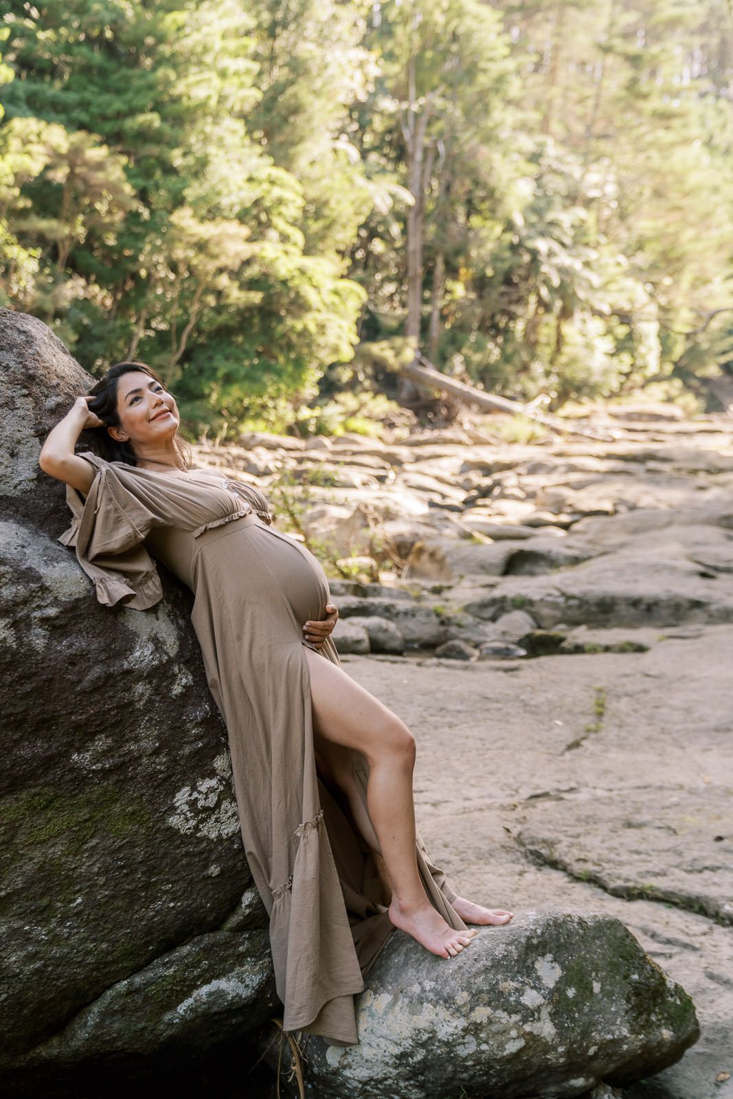 Pregnant woman in tan dress leaning against rock, smiling outdoors in sunlit forest setting.