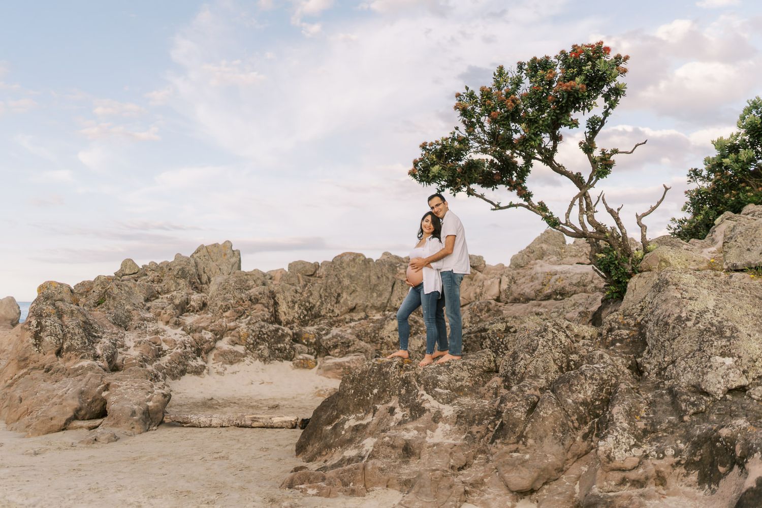 Couple embracing on rocky coastal landscape with windswept tree and cloudy sky backdrop.