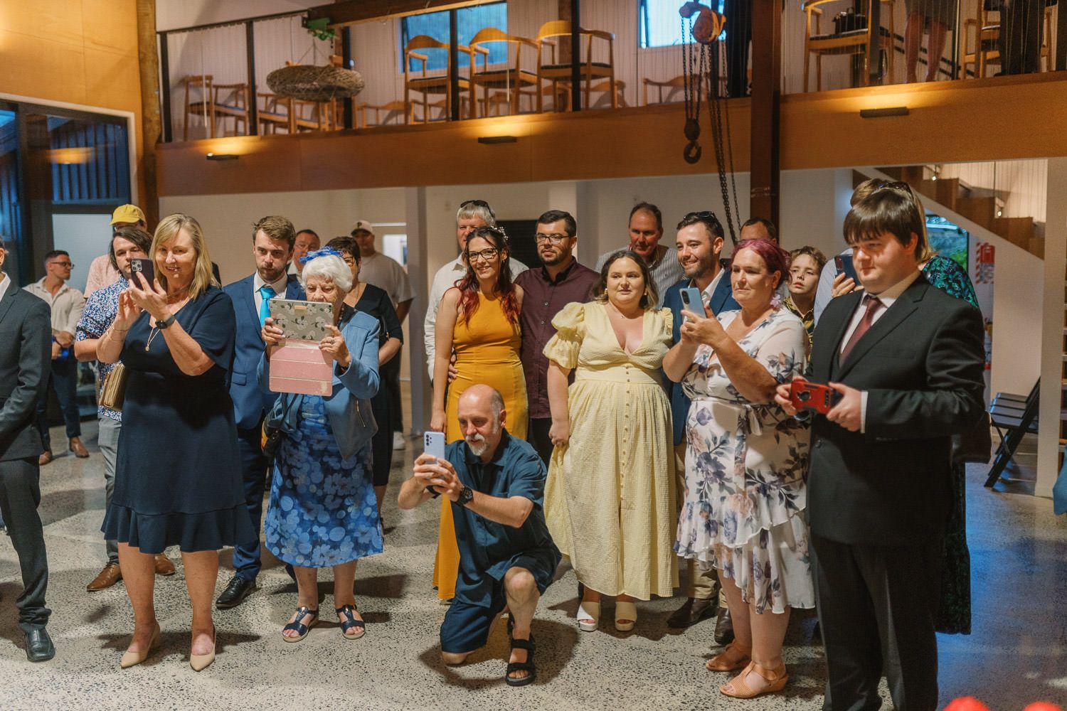 Group of guests smiling and applauding at an indoor wedding reception celebration in a warmly lit venue.