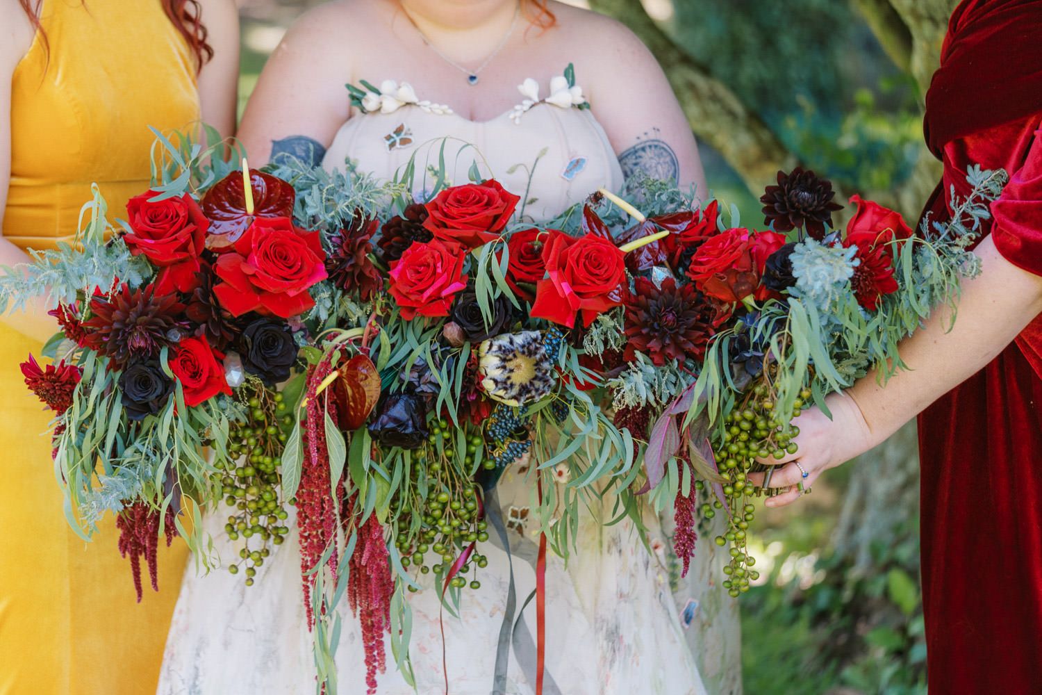 Bride holding a lush cascading bouquet of red roses, dark blooms, and trailing greenery, flanked by bridesmaids in yellow and red.