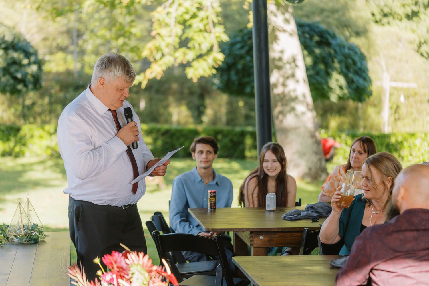 Man giving speech with microphone at outdoor wedding reception while guests sit at wooden table in sunny garden.