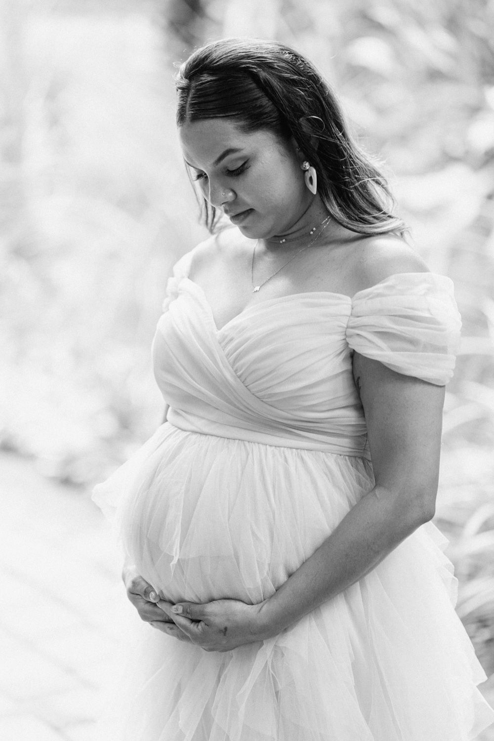 Black and white maternity photo of a pregnant woman in an off-shoulder white tulle gown, gently cradling her baby bump.