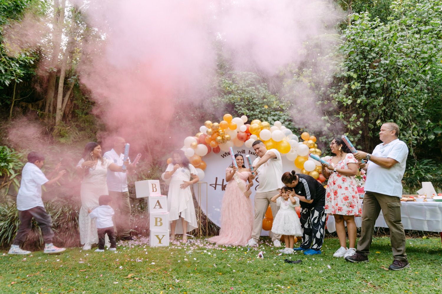 Gender reveal party with pink smoke, balloons, and guests celebrating outdoors in a lush garden setting.