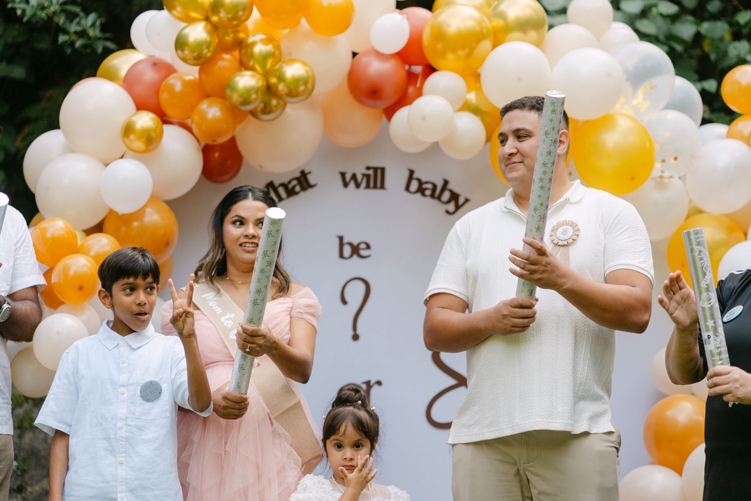 Family at a gender reveal party with balloon arch, holding confetti poppers near a question mark sign.