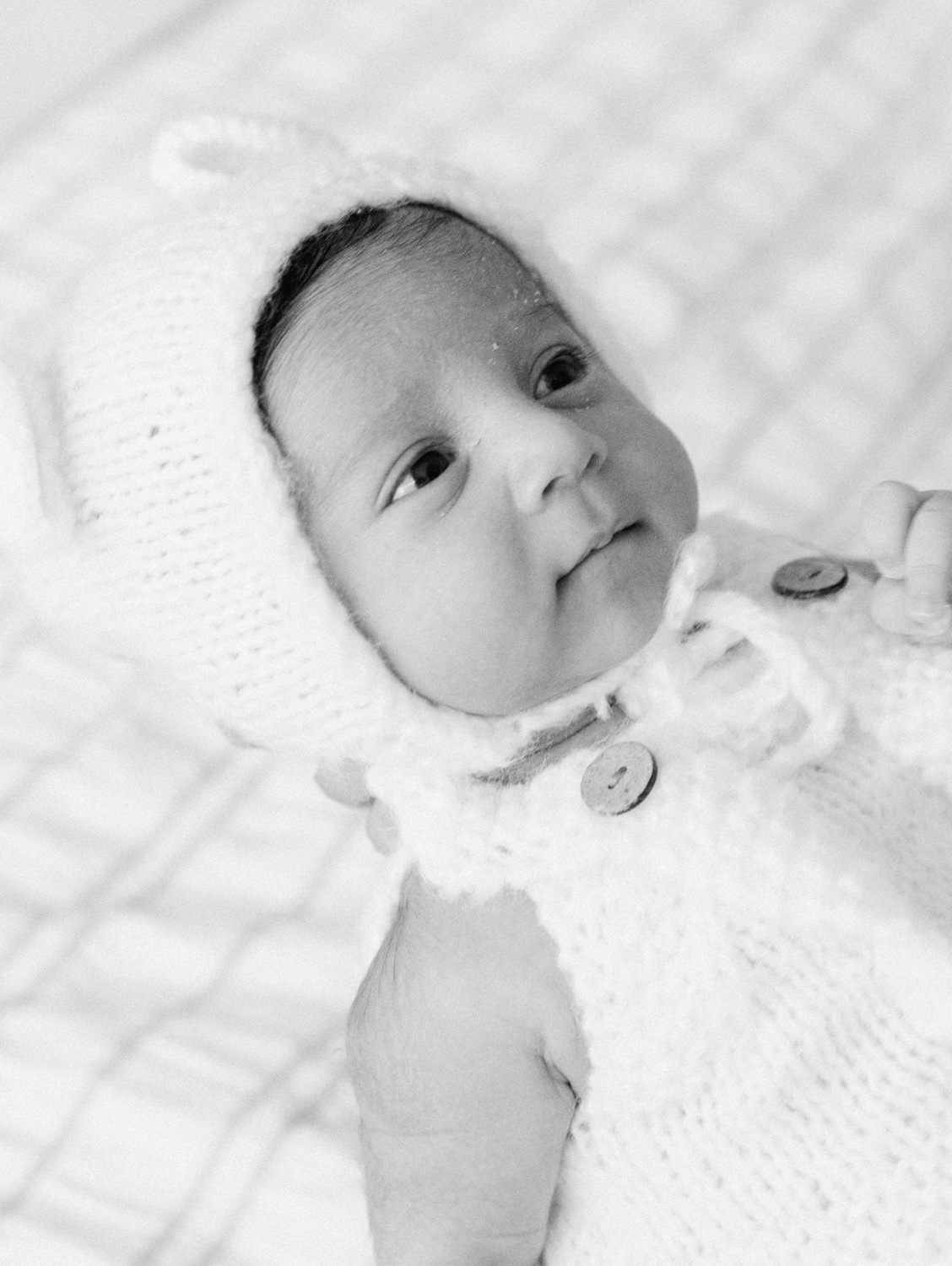 Black and white close-up of newborn baby wearing a white knit bear bonnet and sweater.