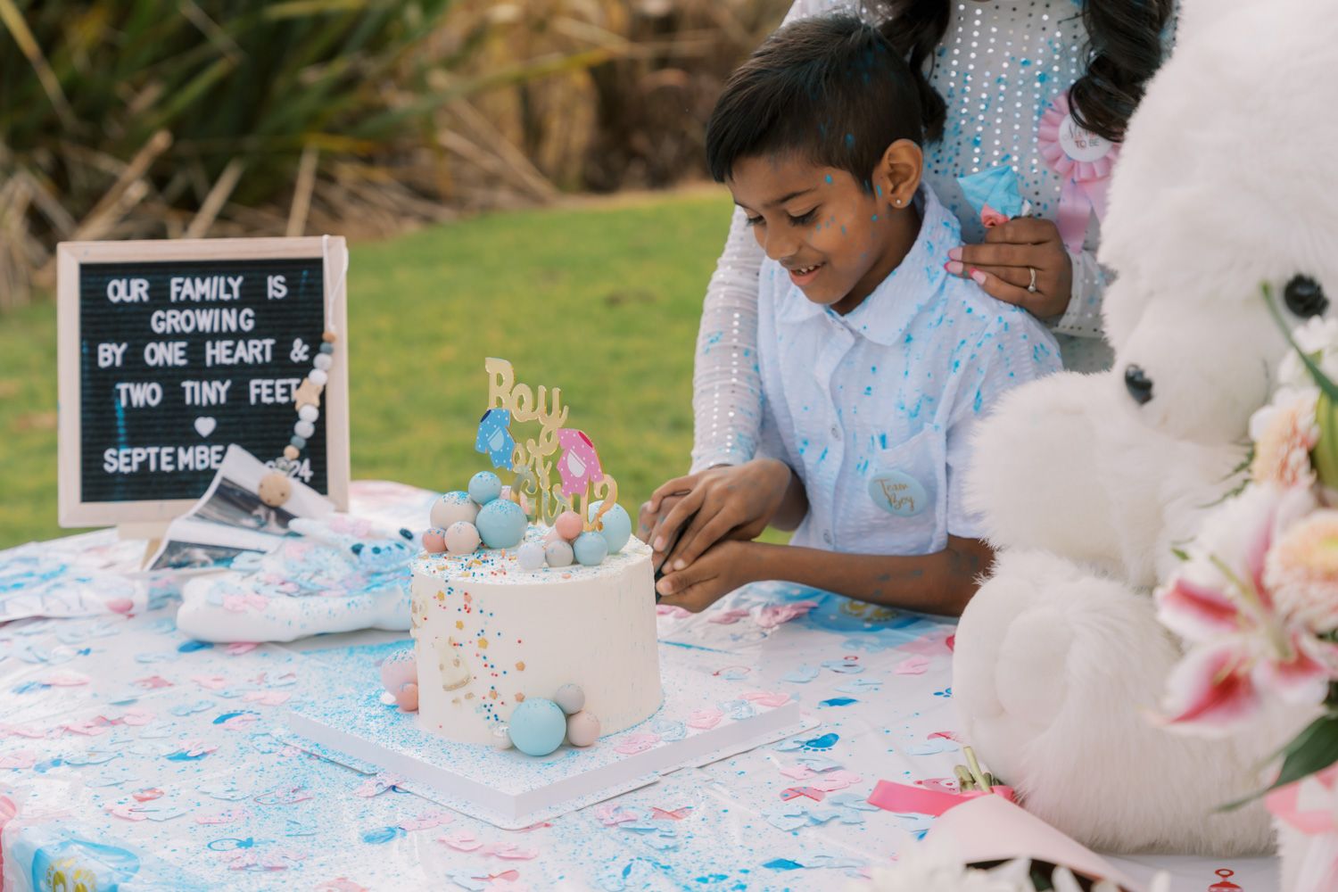 A boy writes at a gender reveal party table with a cake, decorations, and a sign announcing a new baby due in September.