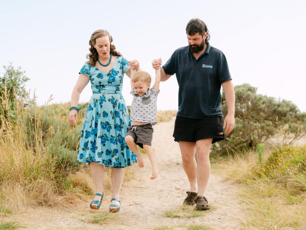 Family of three walking on sandy path, parents holding toddler's hands, woman in floral dress, man in black polo.