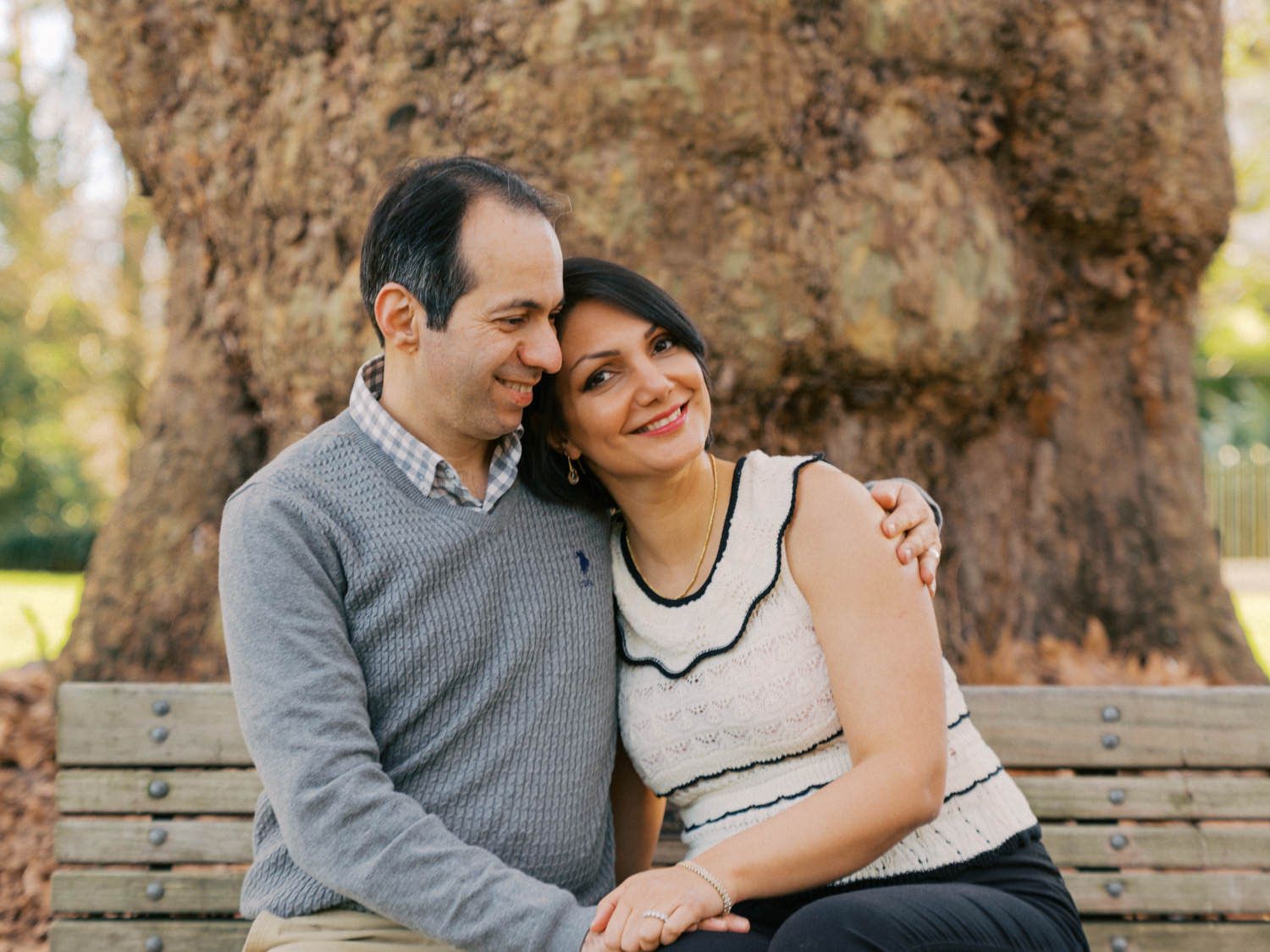 Couple sitting closely on a park bench, smiling warmly, with a large tree in the background.