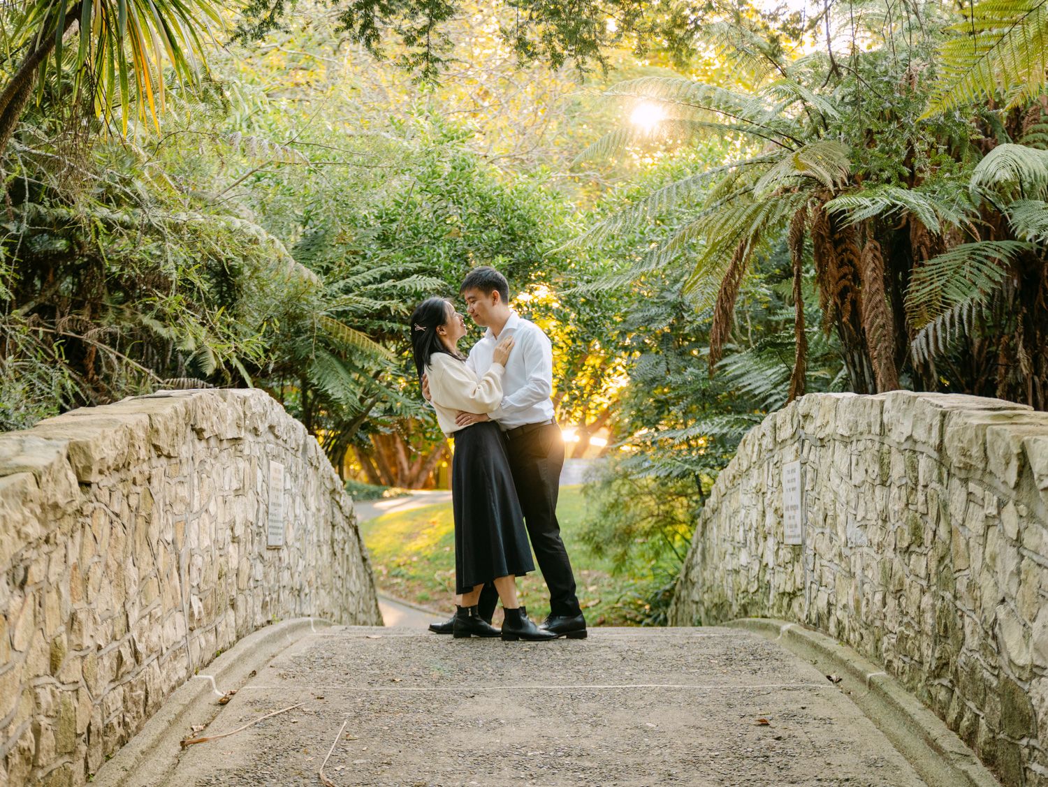 Couple embracing on a stone bridge surrounded by lush tropical ferns and trees with golden sunlight.