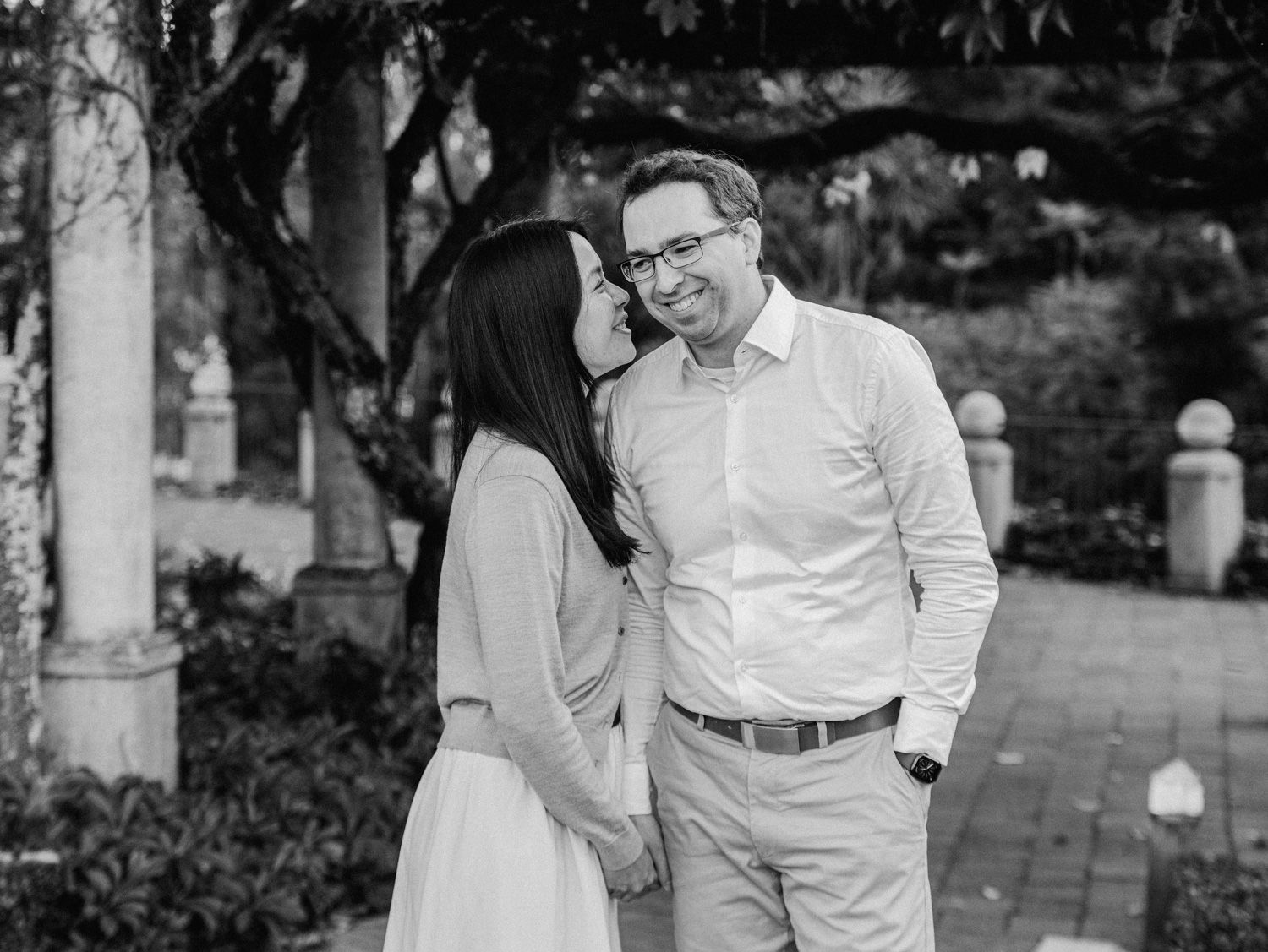 Black and white photo of a smiling couple holding hands in a garden, sharing a romantic moment outdoors.