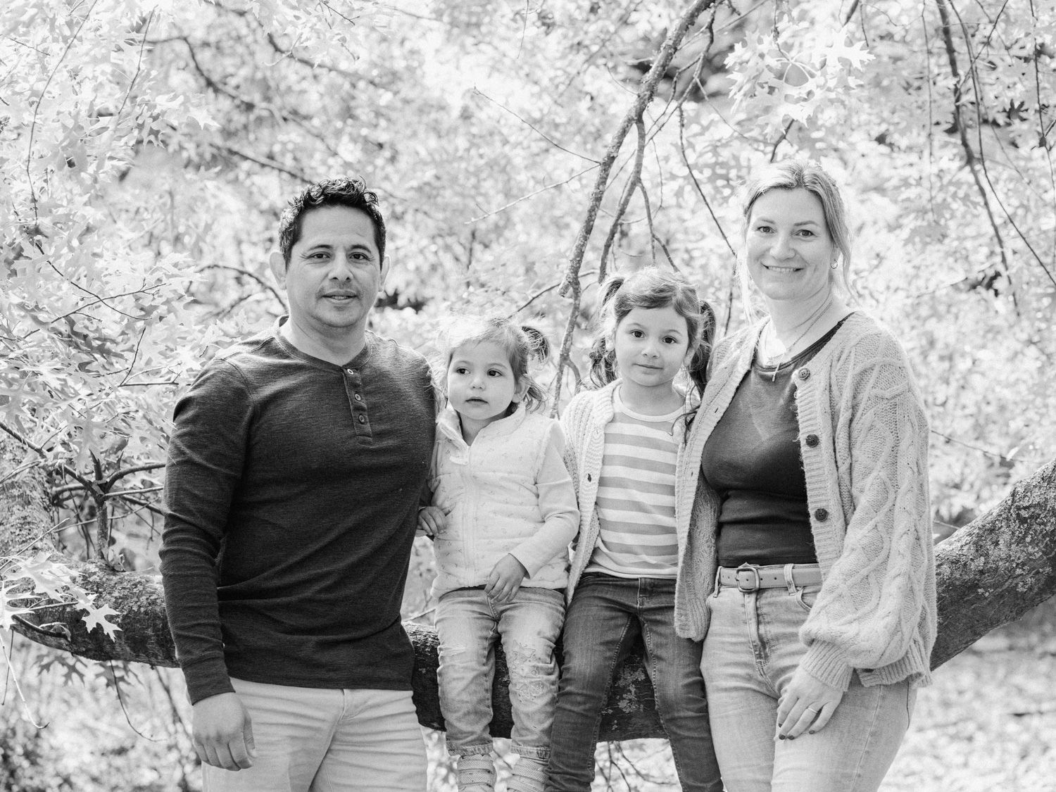 Black and white family portrait of parents with two young children outdoors among trees.