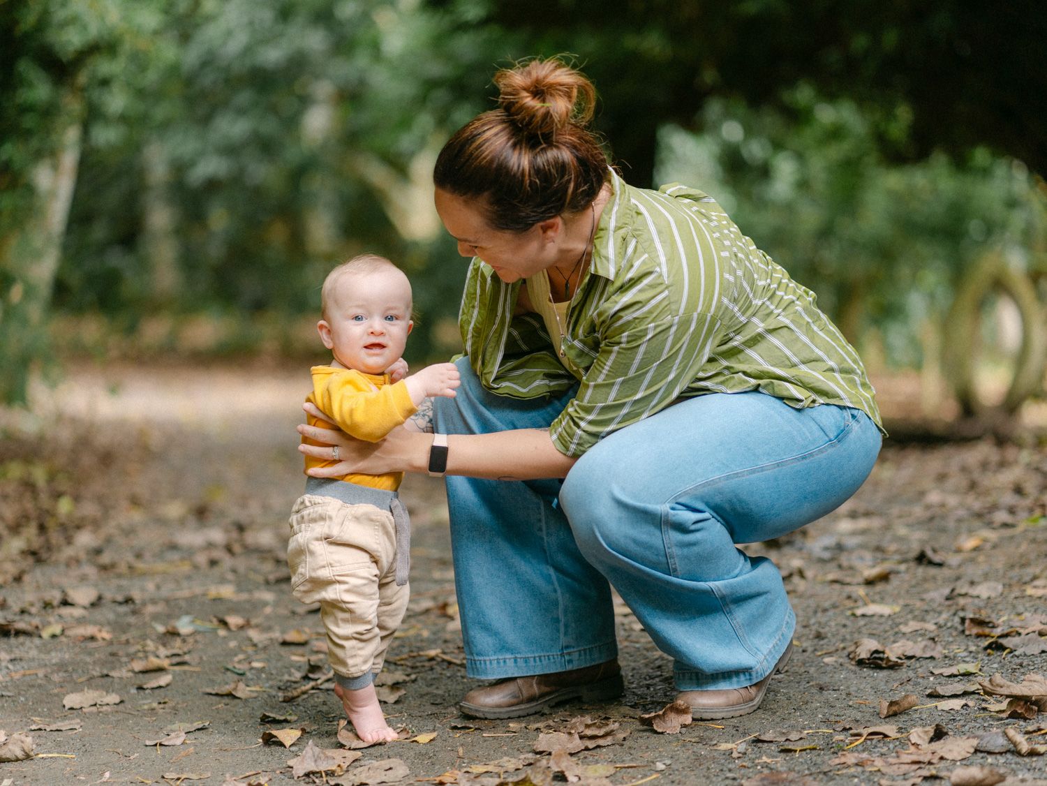 A mother crouches down helping her baby take first steps on a leaf-covered path in a lush green park.