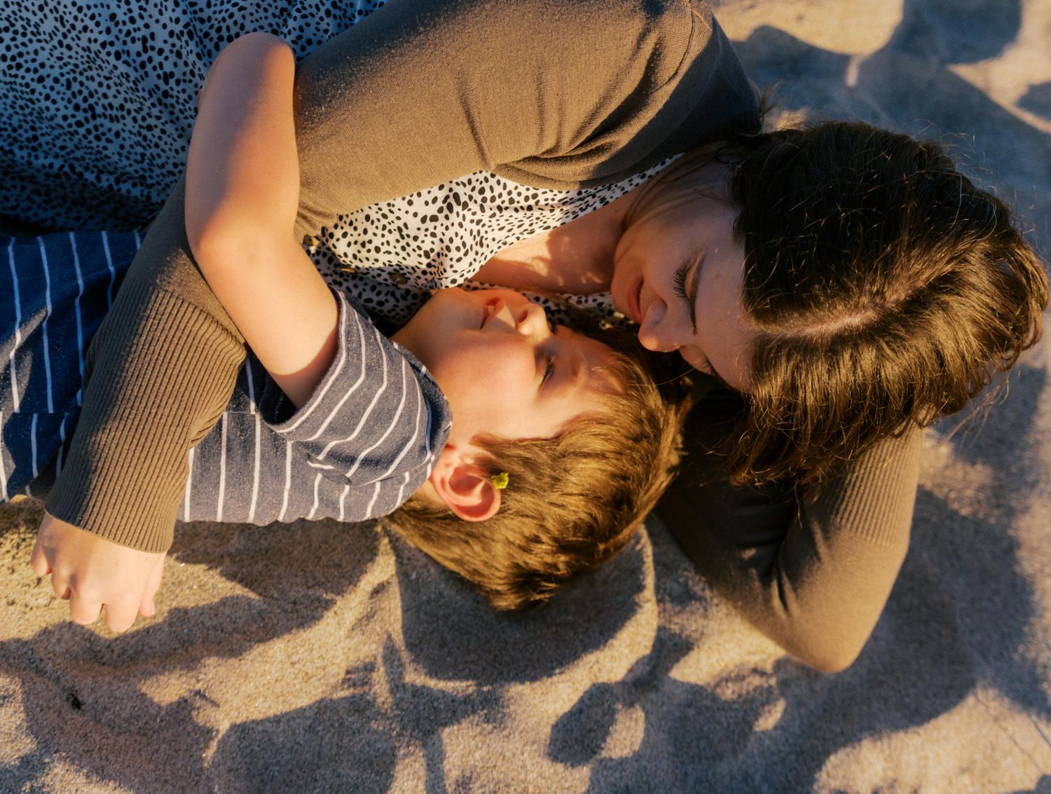 A mother tenderly kisses her young child while lying together on sandy beach in warm golden sunlight.