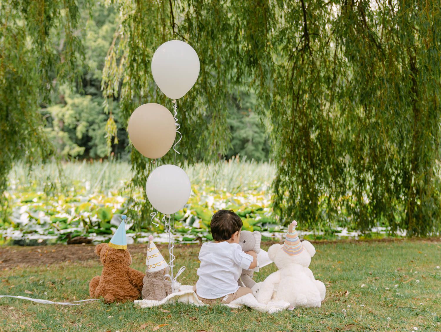 Baby sitting on grass under a willow tree with white balloons and stuffed animals at a first birthday party.