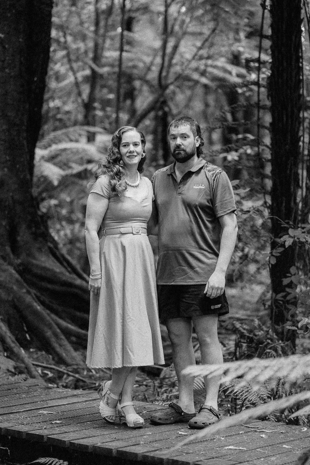 Black and white photo of a couple standing on a wooden boardwalk in a lush forest setting.