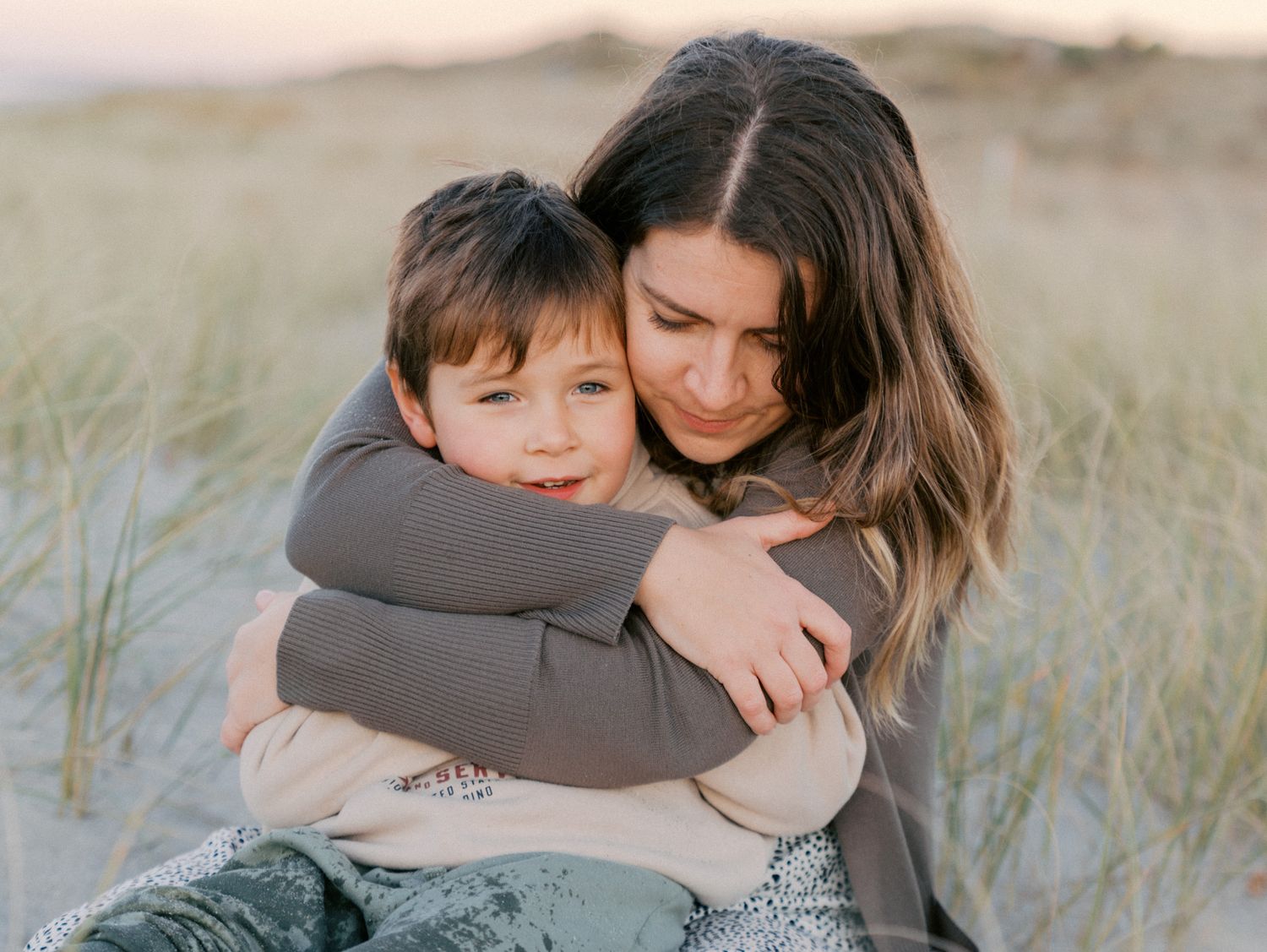 A mother tenderly hugs her young son on a sandy beach at sunset, surrounded by dune grass.