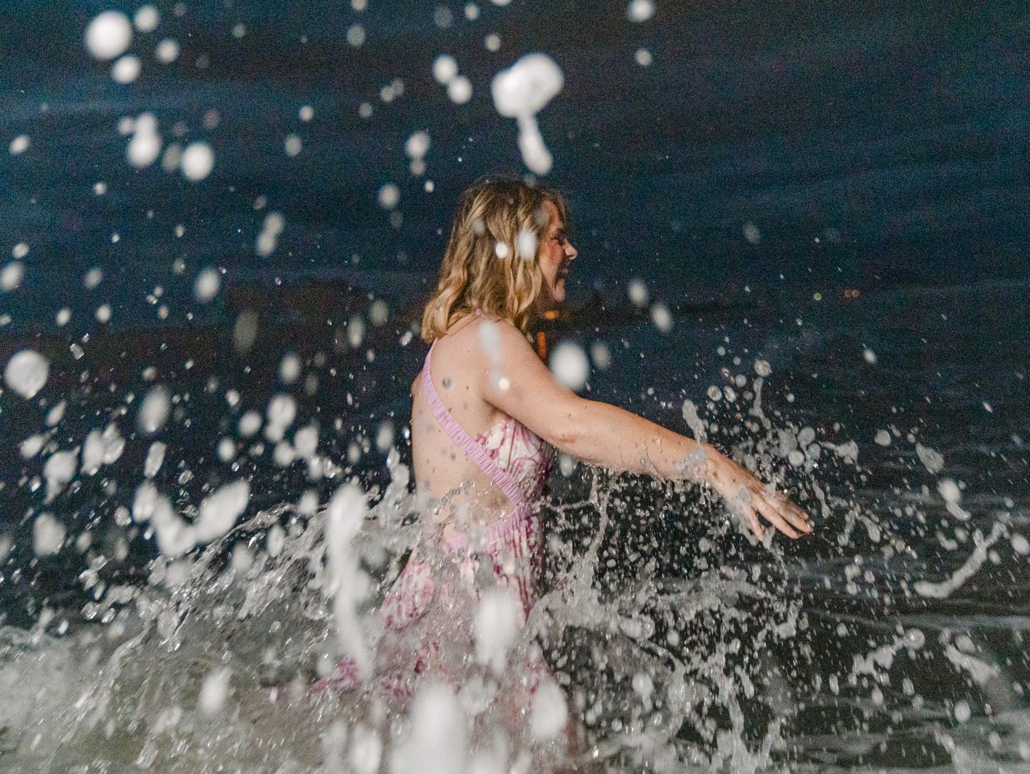 Woman in pink swimsuit splashing in ocean waves at night, water droplets frozen in motion around her.