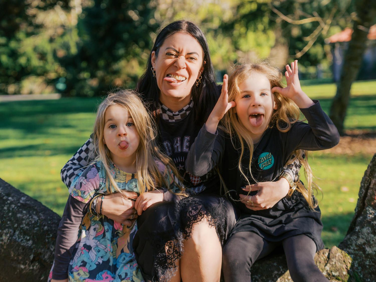 Woman and two young girls making funny faces together while sitting on a rock in a park.