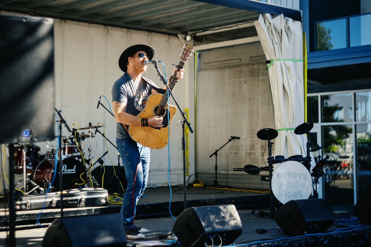 Musician playing acoustic guitar on outdoor stage wearing black hat and jeans at a live music event.