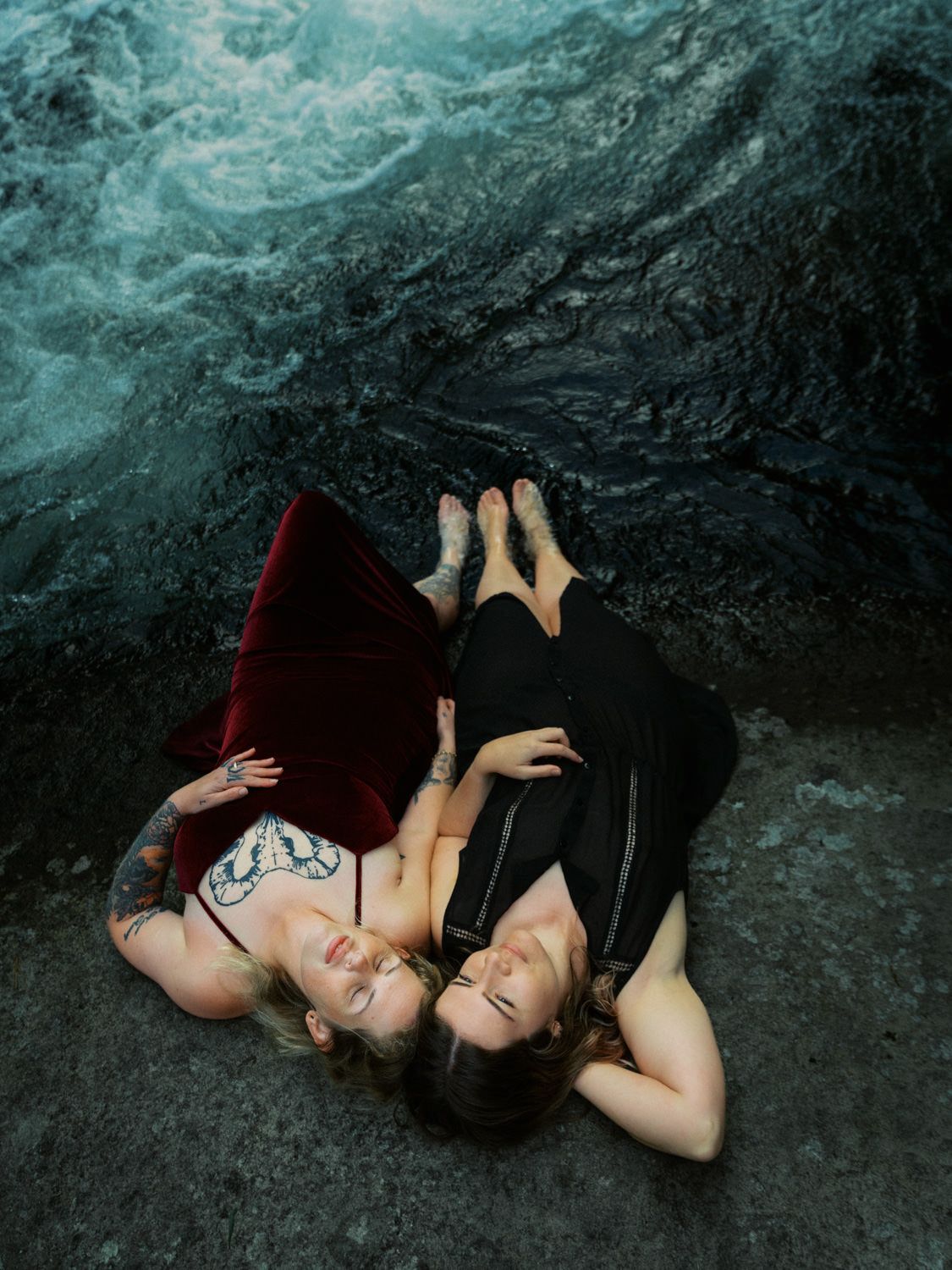 Two women in elegant red and black dresses lying on dark rocks near turbulent ocean waves.