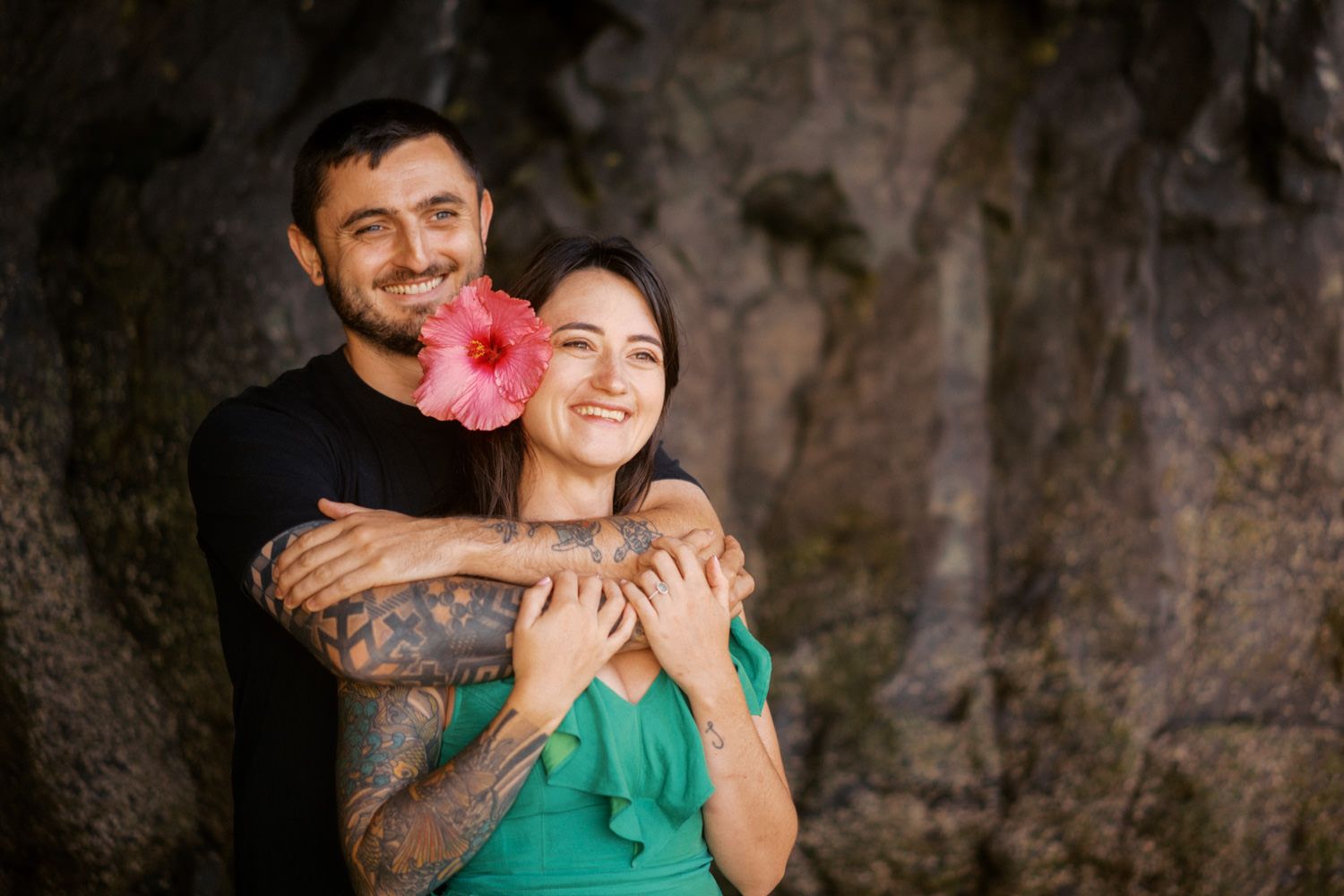 A tattooed man embraces a smiling woman holding a pink hibiscus flower, posing before a rocky backdrop.