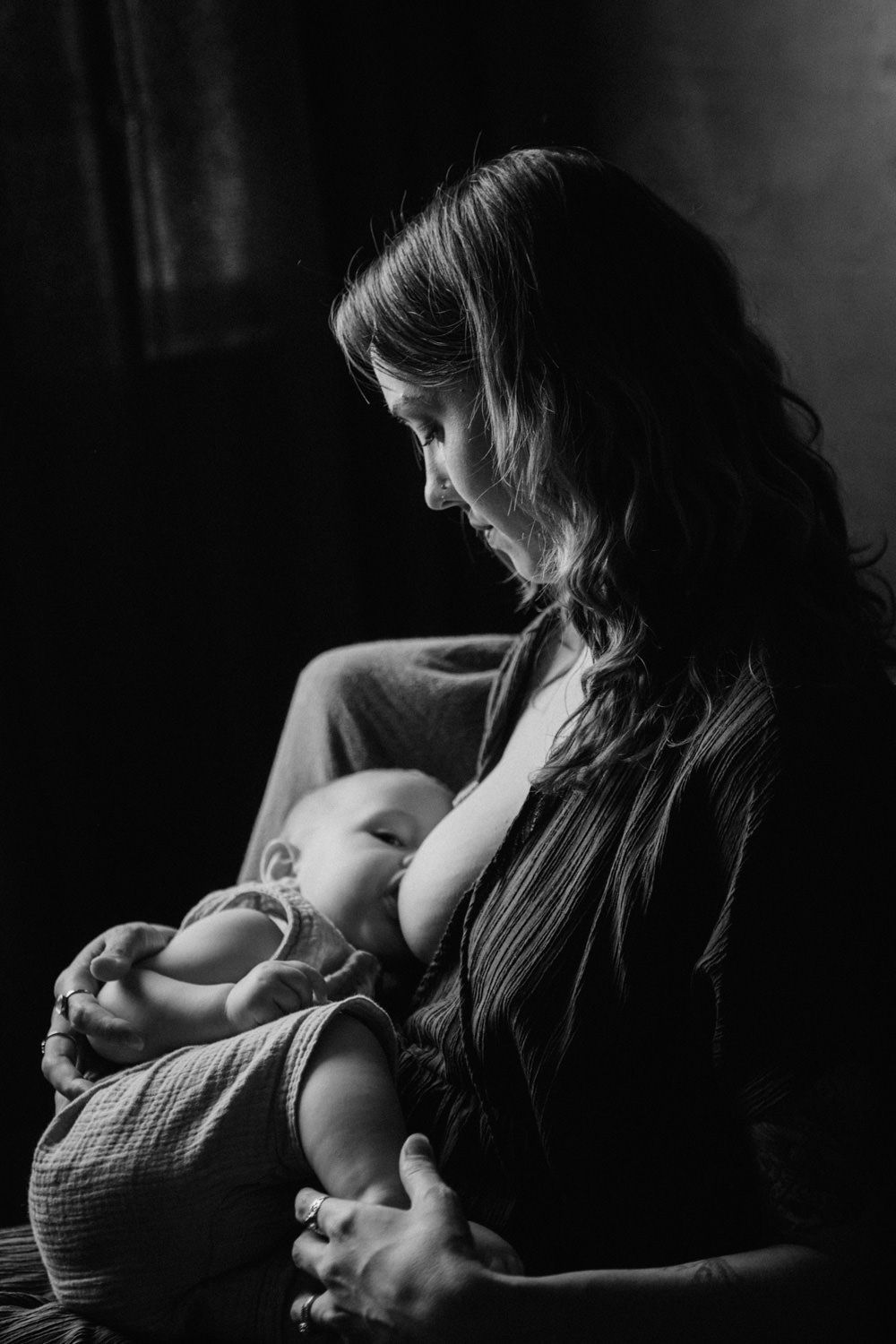 Black and white photo of a mother tenderly breastfeeding her newborn baby in a softly lit room.