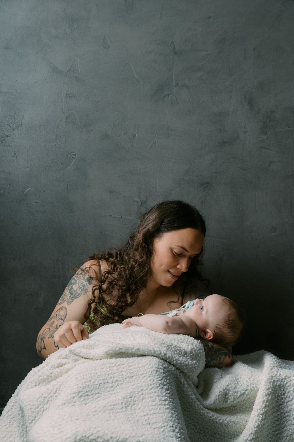 Tattooed mother tenderly gazes at her newborn baby wrapped in a white blanket against a gray backdrop.