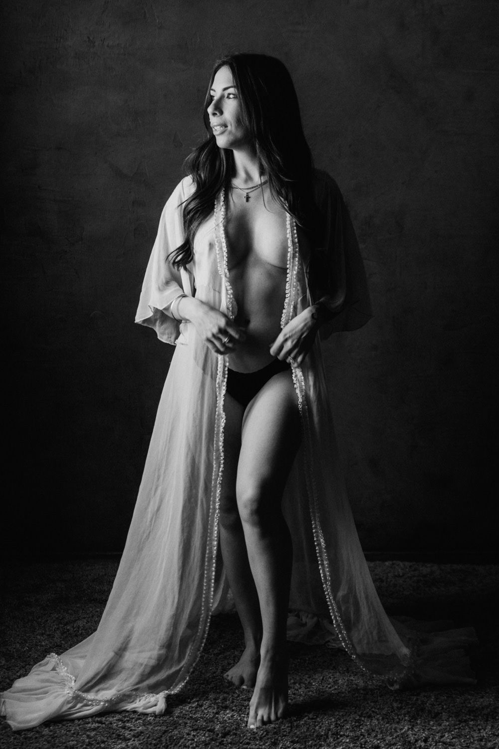 Black and white fine art portrait of a woman in a sheer robe posing elegantly in a studio setting.