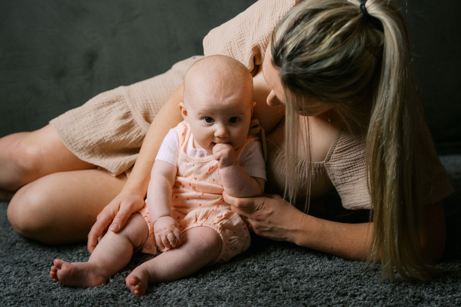 A loving mother sits on a gray carpet, gently kissing her adorable baby dressed in a pink outfit.