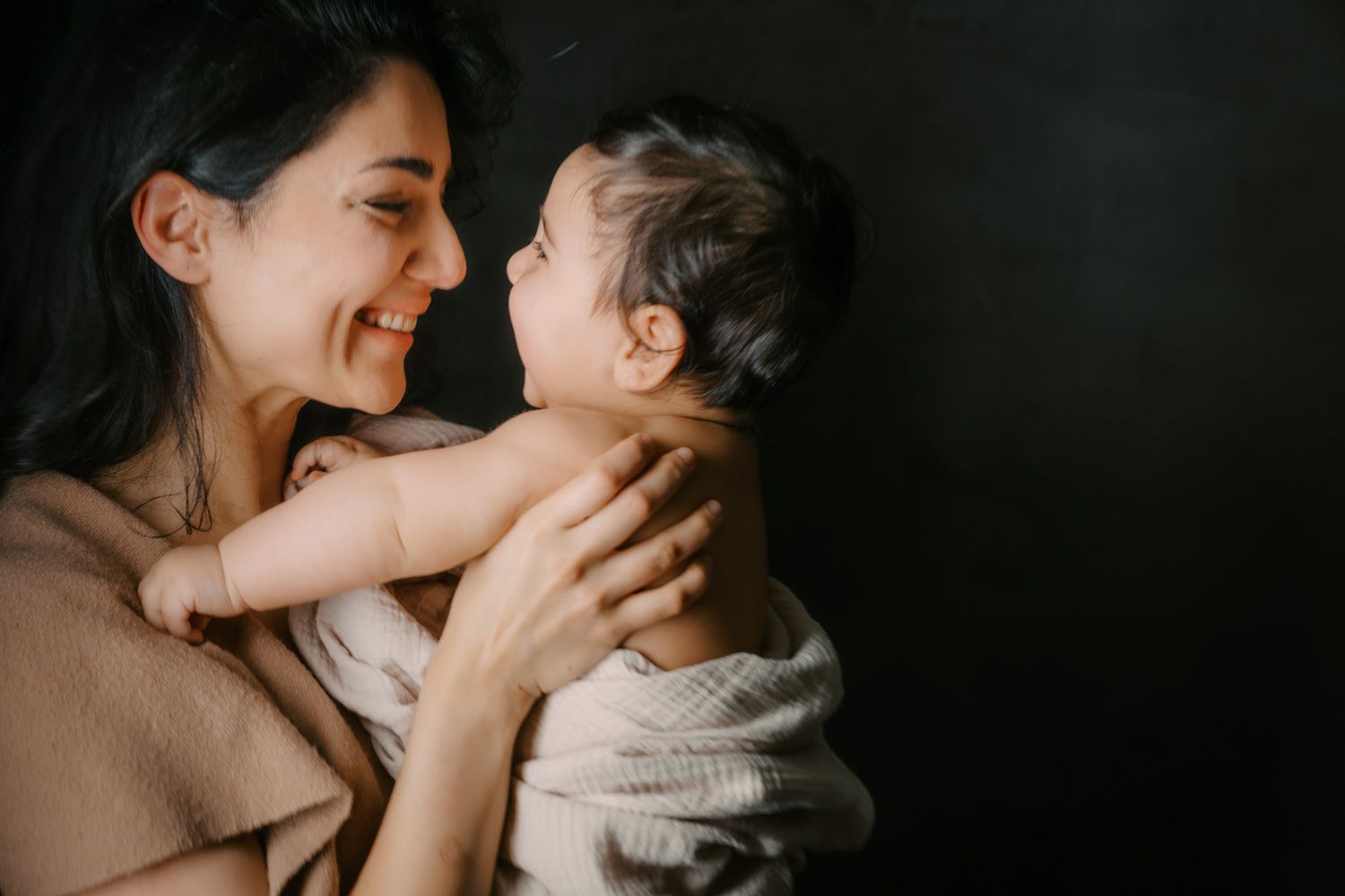A smiling mother tenderly holds her newborn baby, sharing a loving nose-to-nose moment against a dark background.
