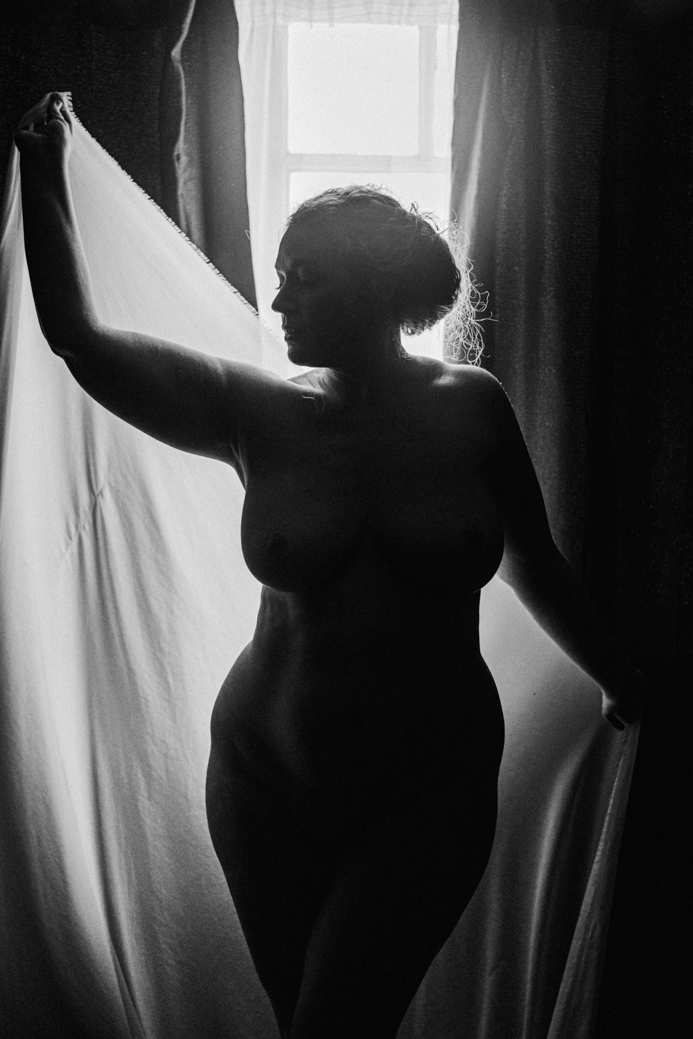 Black and white silhouette of a woman standing by a window with flowing curtains in artistic boudoir photography.