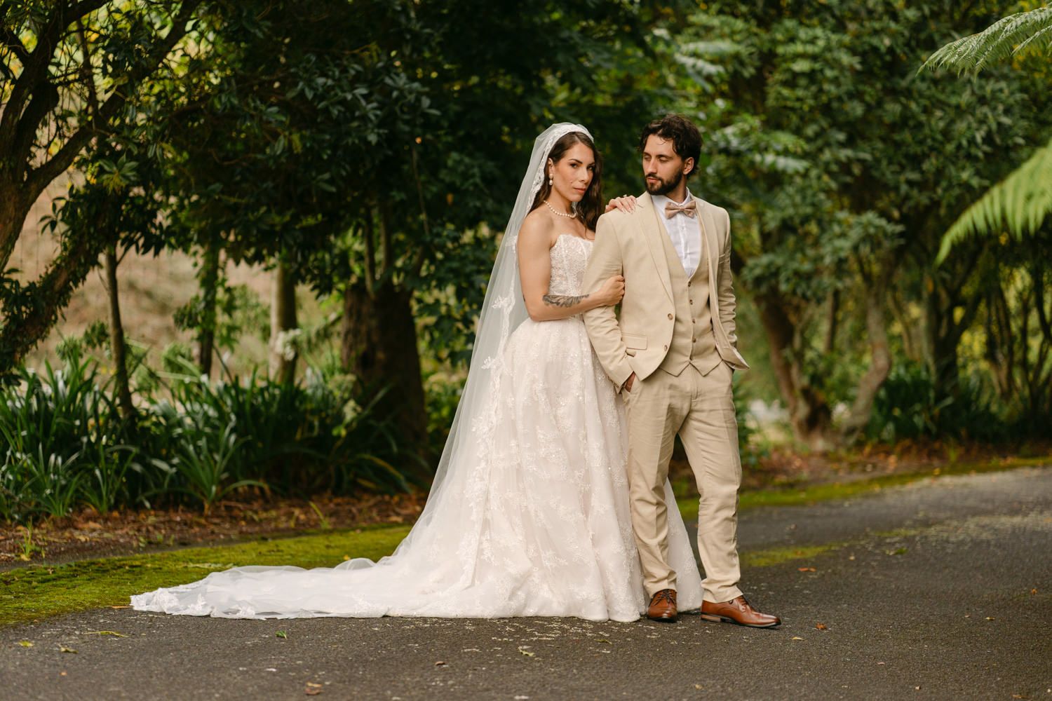 Bride in white gown with veil and groom in tan suit pose together on a tree-lined path at outdoor wedding.