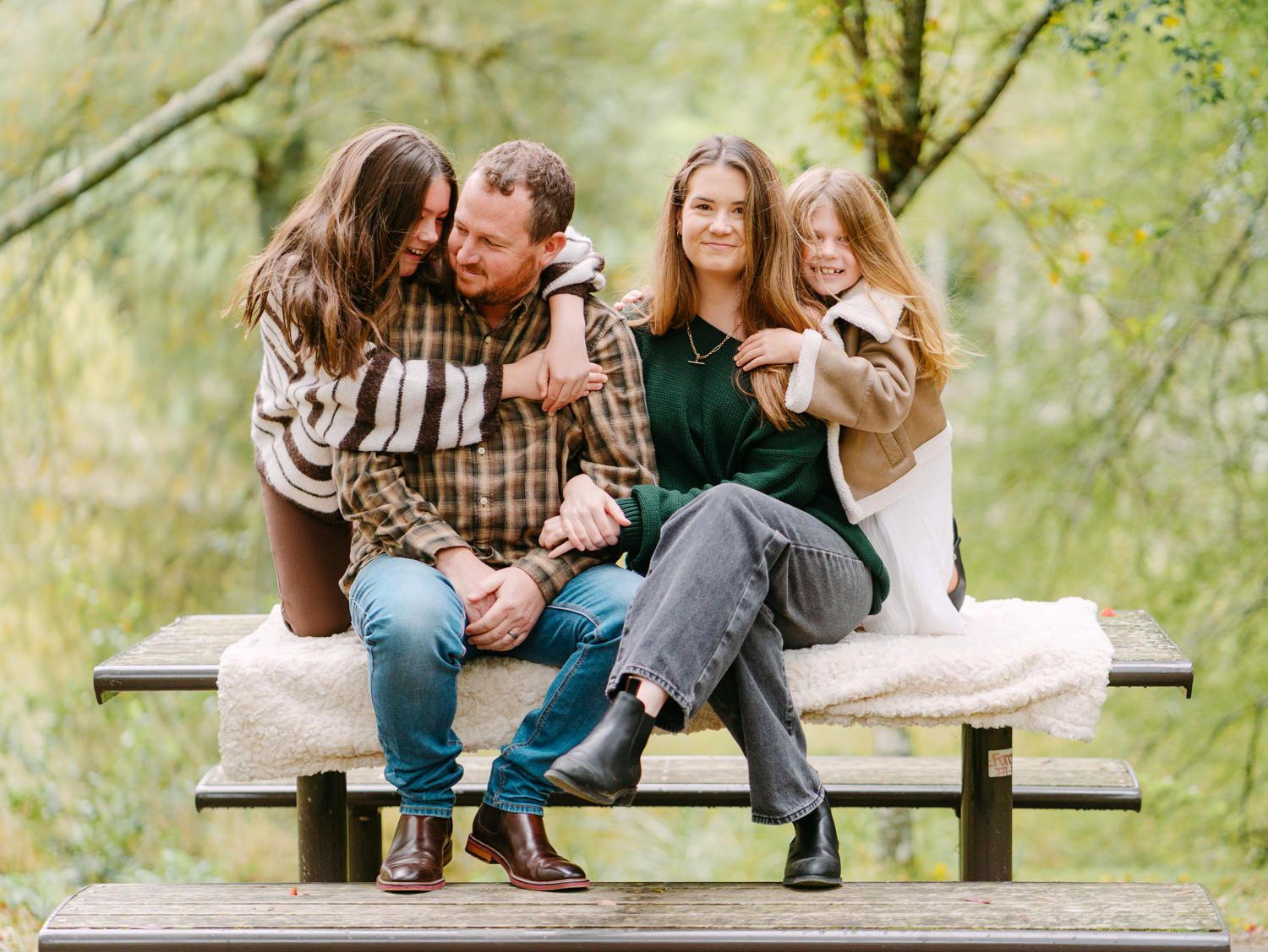 Family of four smiling and embracing on a wooden bench surrounded by lush green trees in an outdoor setting.