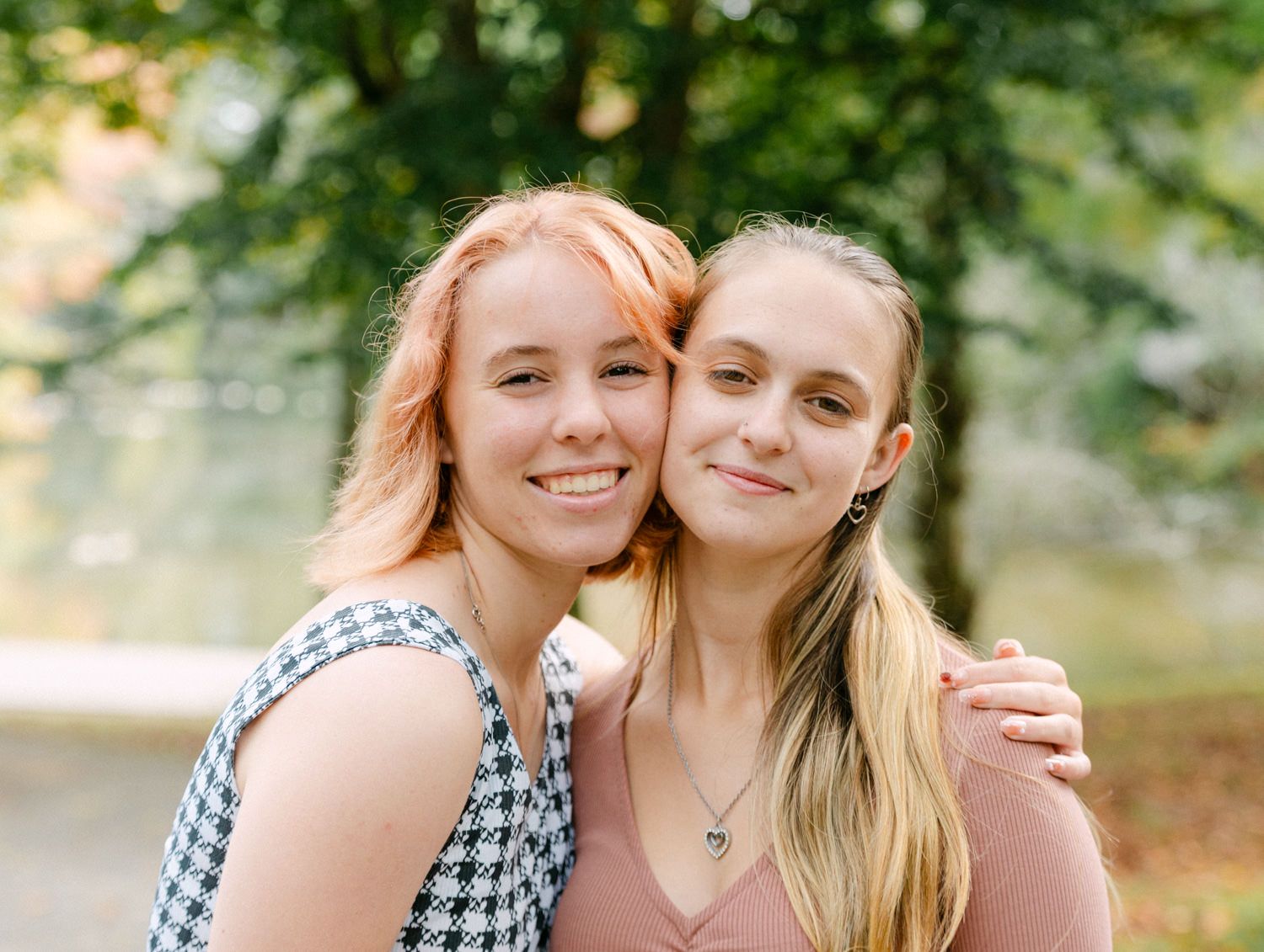 Two smiling young women with blonde hair pose together outdoors in a park setting with green trees in the background.