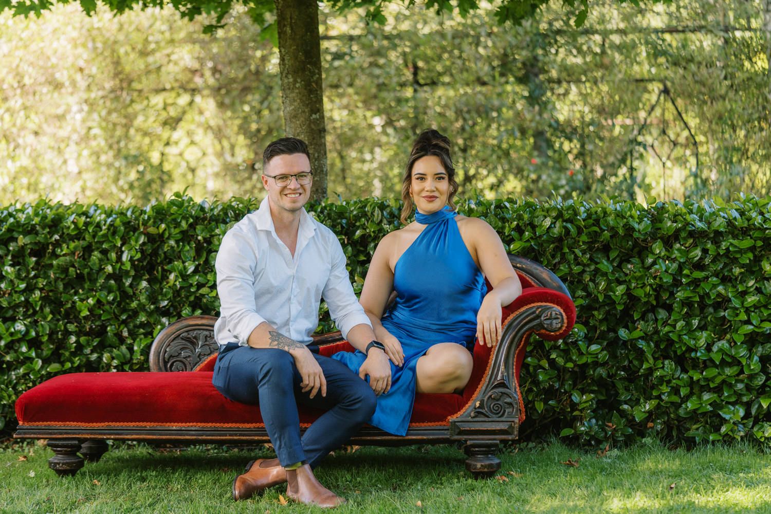 A smiling couple sits on a ornate red velvet chaise lounge outdoors, man in white shirt and woman in blue dress.