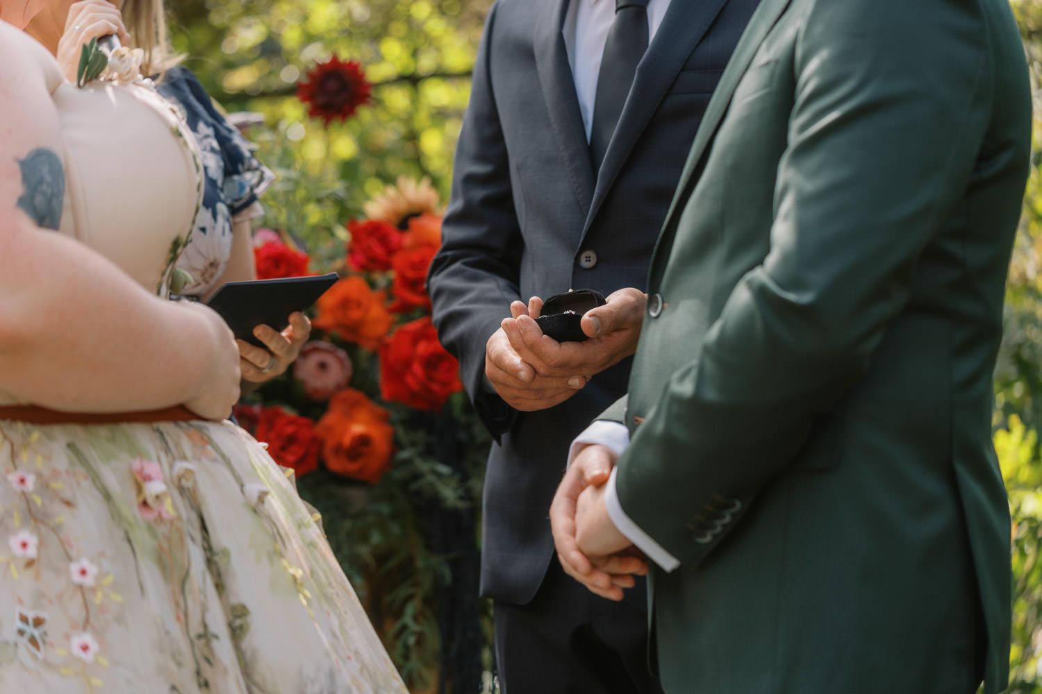 Couple exchanging rings at outdoor wedding ceremony with vibrant red and orange floral arrangements in background.